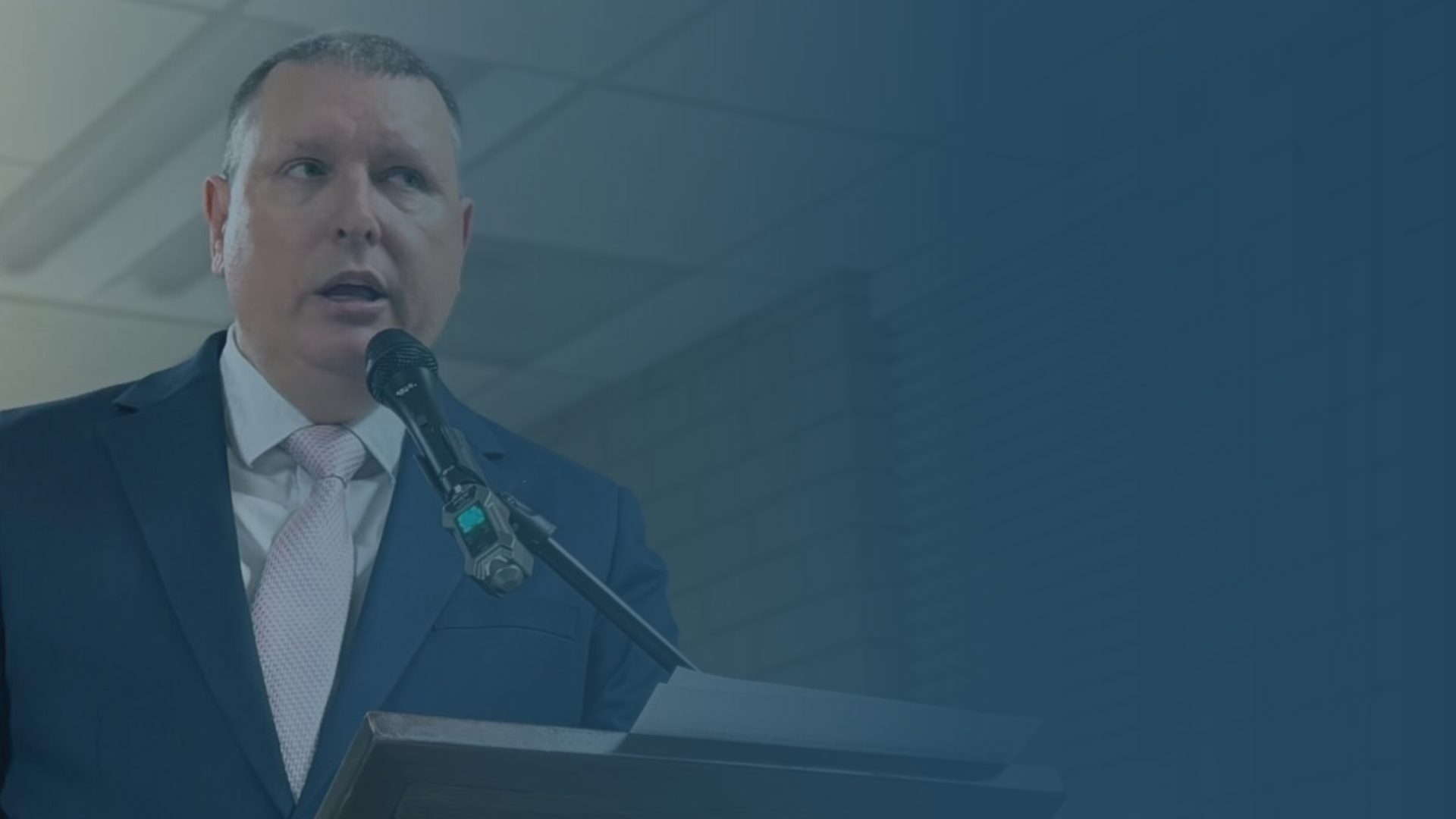 Shane Lewis in a suit speaking at a podium with a microphone, in an indoor setting.