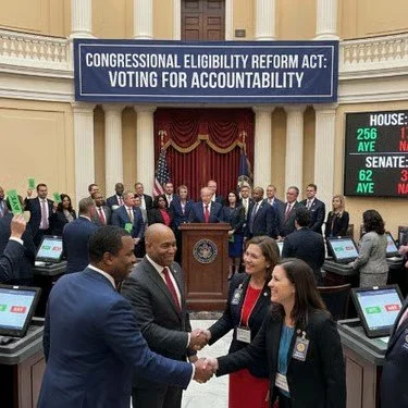 A group of lawmakers in a legislative chamber, with some shaking hands and others standing around, beneath a sign that reads 'Congressional Eligibility Reform Act: Voting for Accountability' on a digital display.