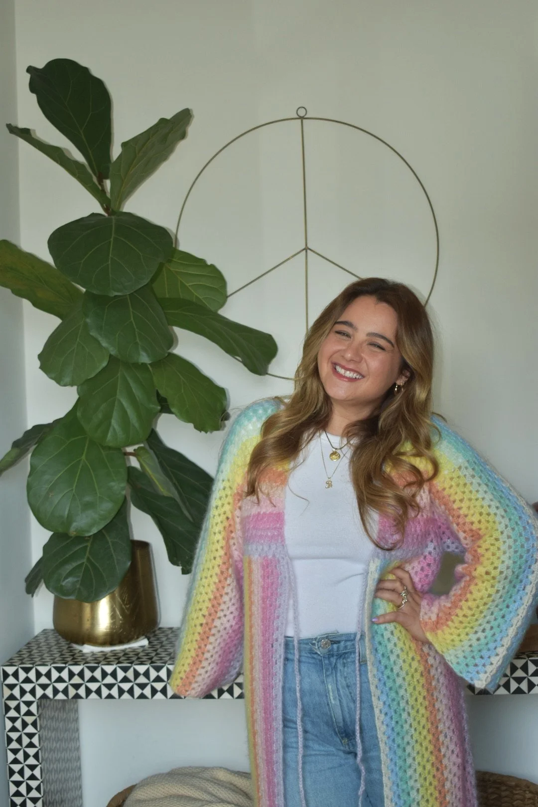 A smiling woman with long wavy hair wearing a colorful striped crochet cardigan over a white shirt, standing indoors next to a large green leafy plant and a black and white patterned table with a gold pot. A peace sign art hangs on the white wall behind her.