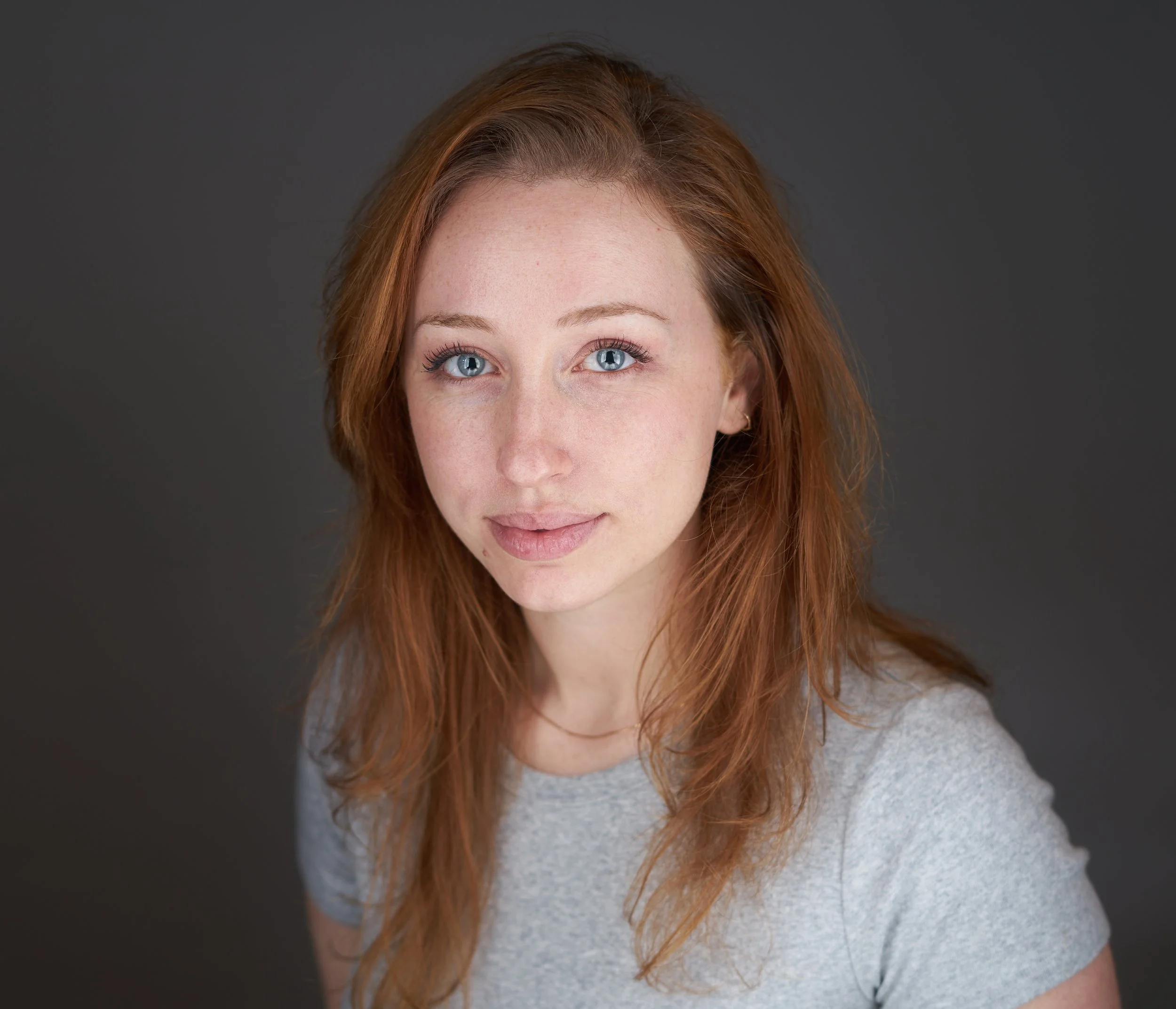 A young woman with red hair and blue eyes looking at the camera, wearing a gray t-shirt, against a dark gray background.