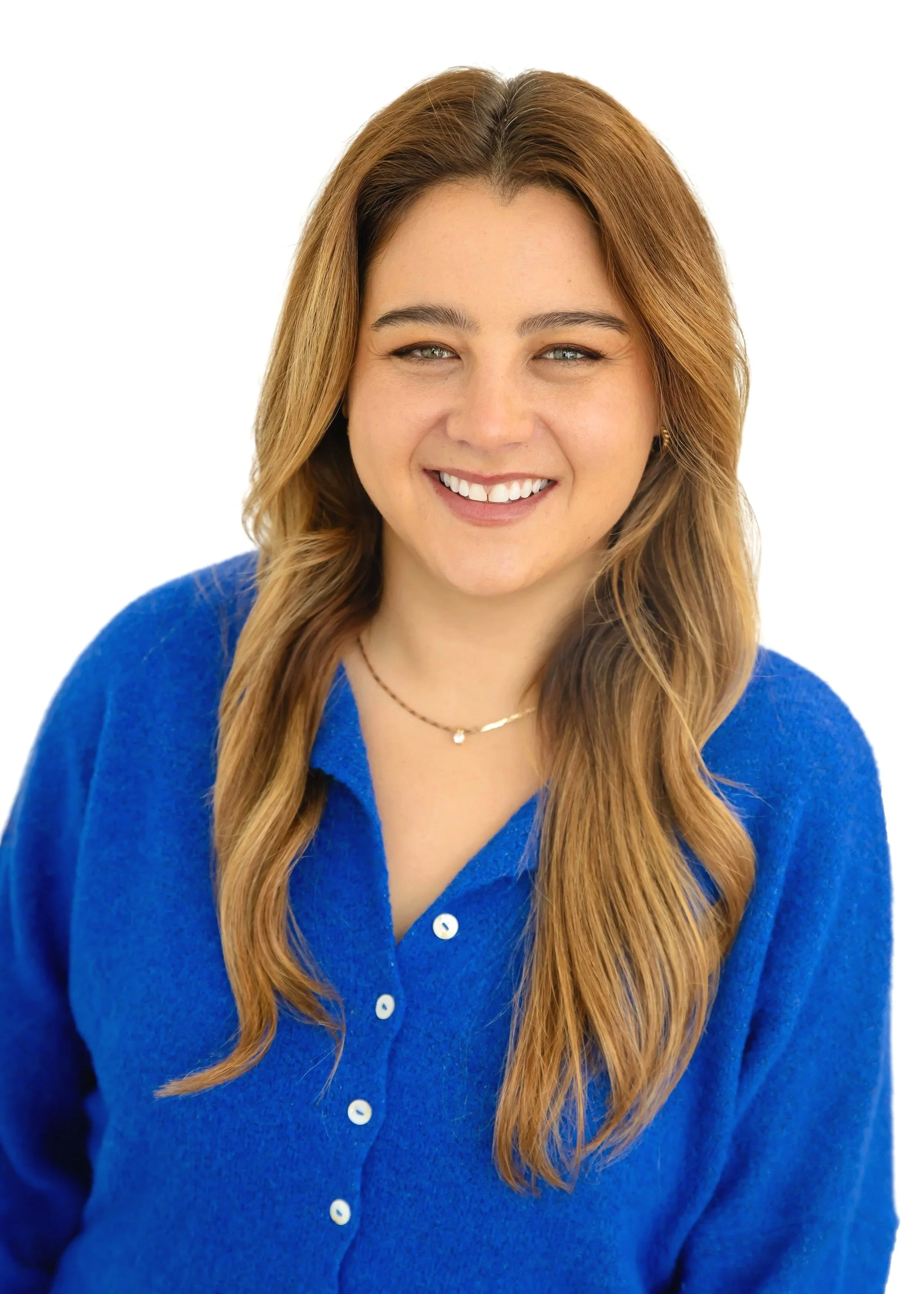 A young woman with long, wavy, light brown hair smiling at the camera, wearing a bright blue button-up sweater and a delicate necklace against a plain white background.