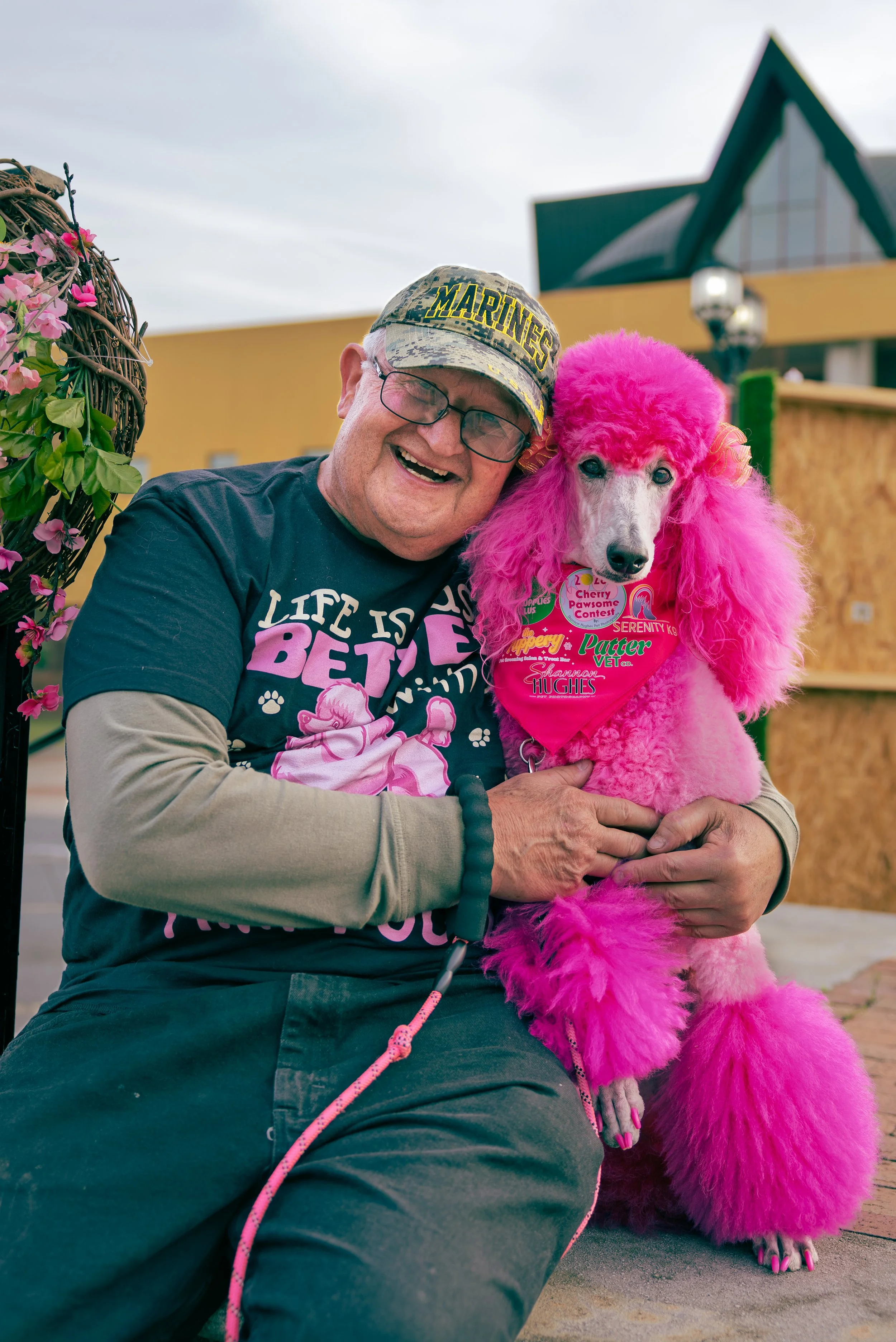 A man with glasses, wearing a camouflage cap and a dark T-shirt, smiling while holding a pink poodle dressed in a pink costume with a bandana at a pet paw contest. They are outdoors with a yellow building and cloudy sky in the background.