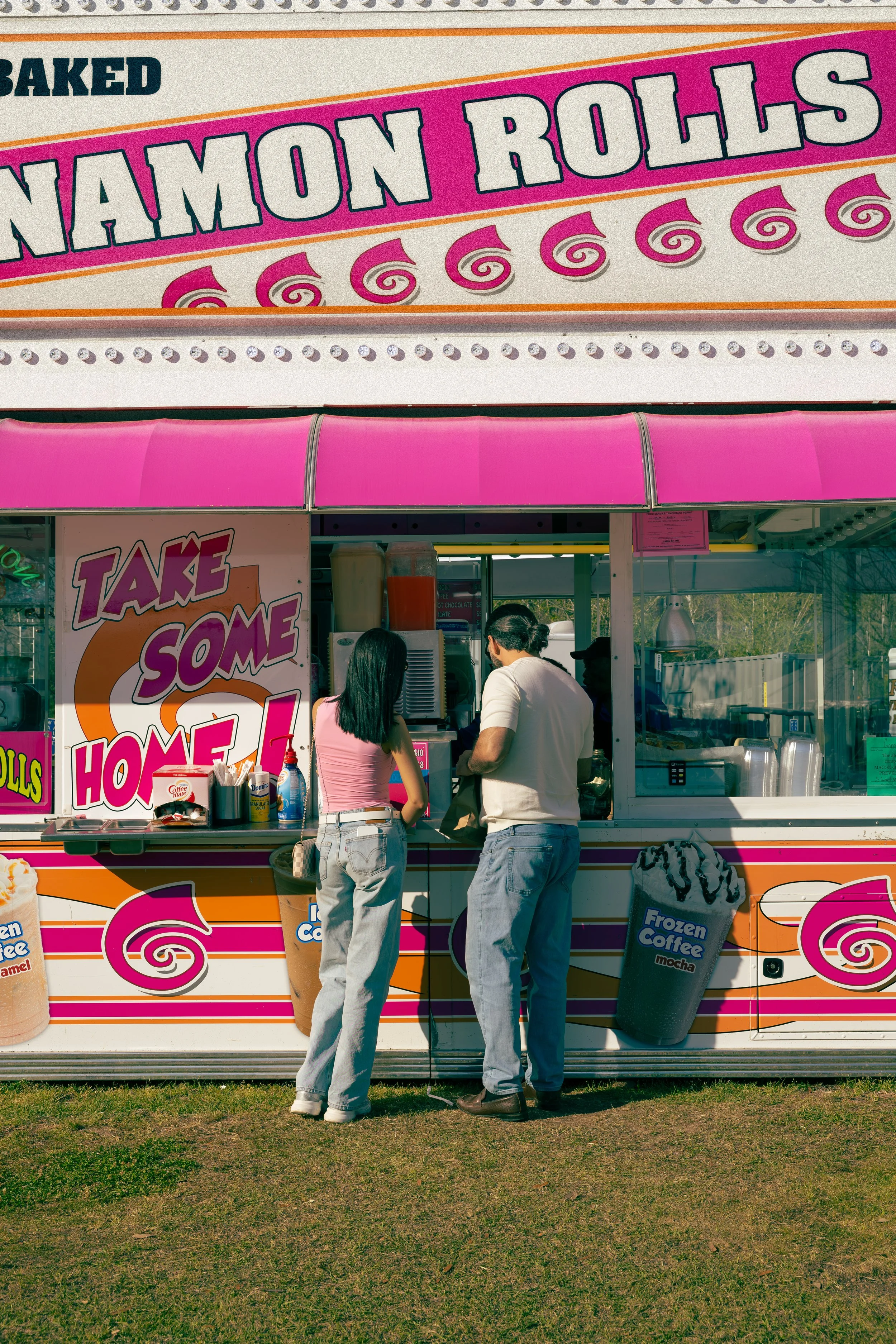 Two people standing in front of a pink and orange shaved ice stand with a large sign reading "Baked Cinnamon Rolls". One woman is wearing a pink top and light jeans, and one man is wearing a white shirt and blue jeans. They are ordering or paying at 