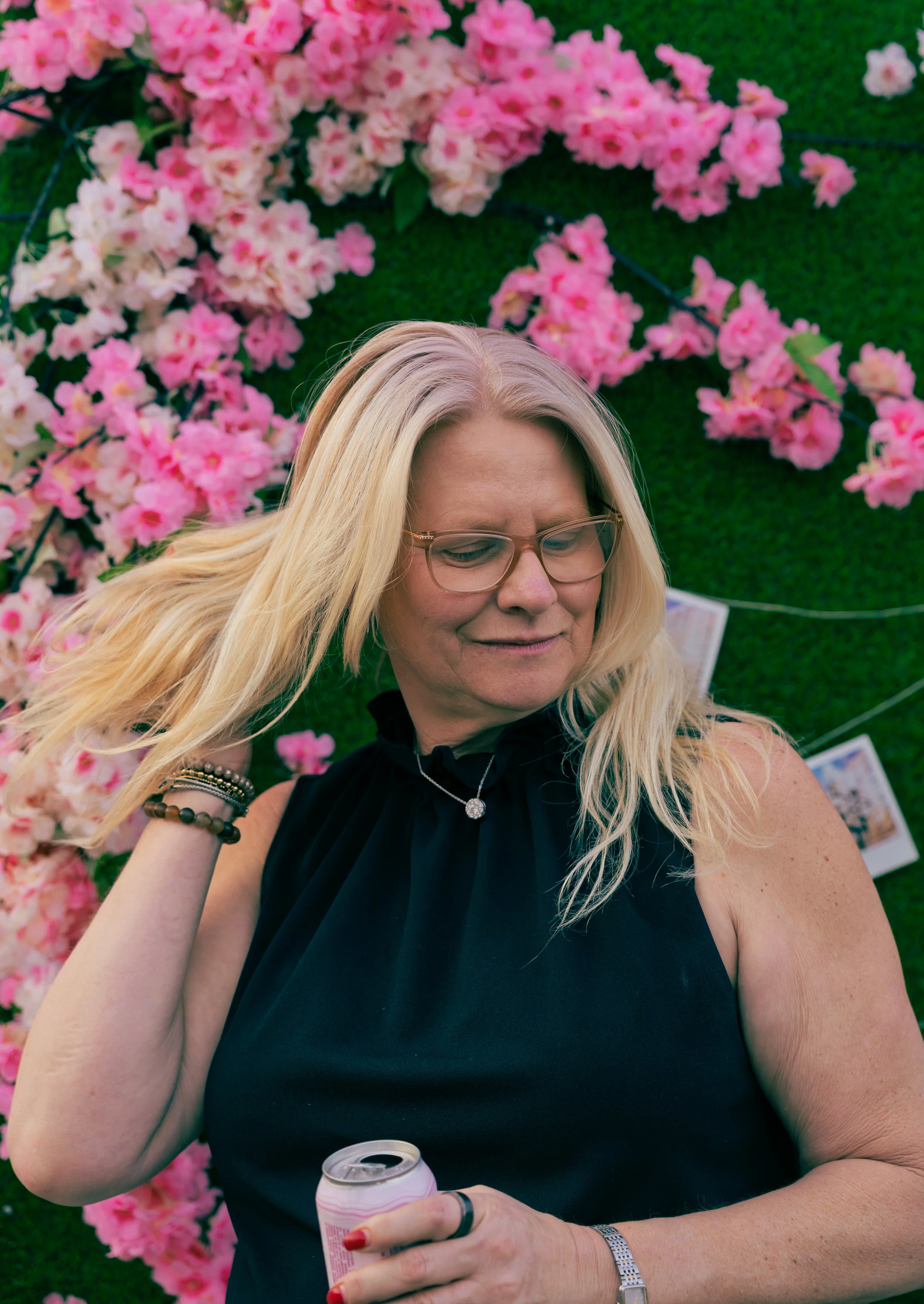 A woman with blonde hair wearing glasses, a black sleeveless top, a necklace, and bracelets, holding a can of soda, standing in front of pink flowers and a green backdrop.
