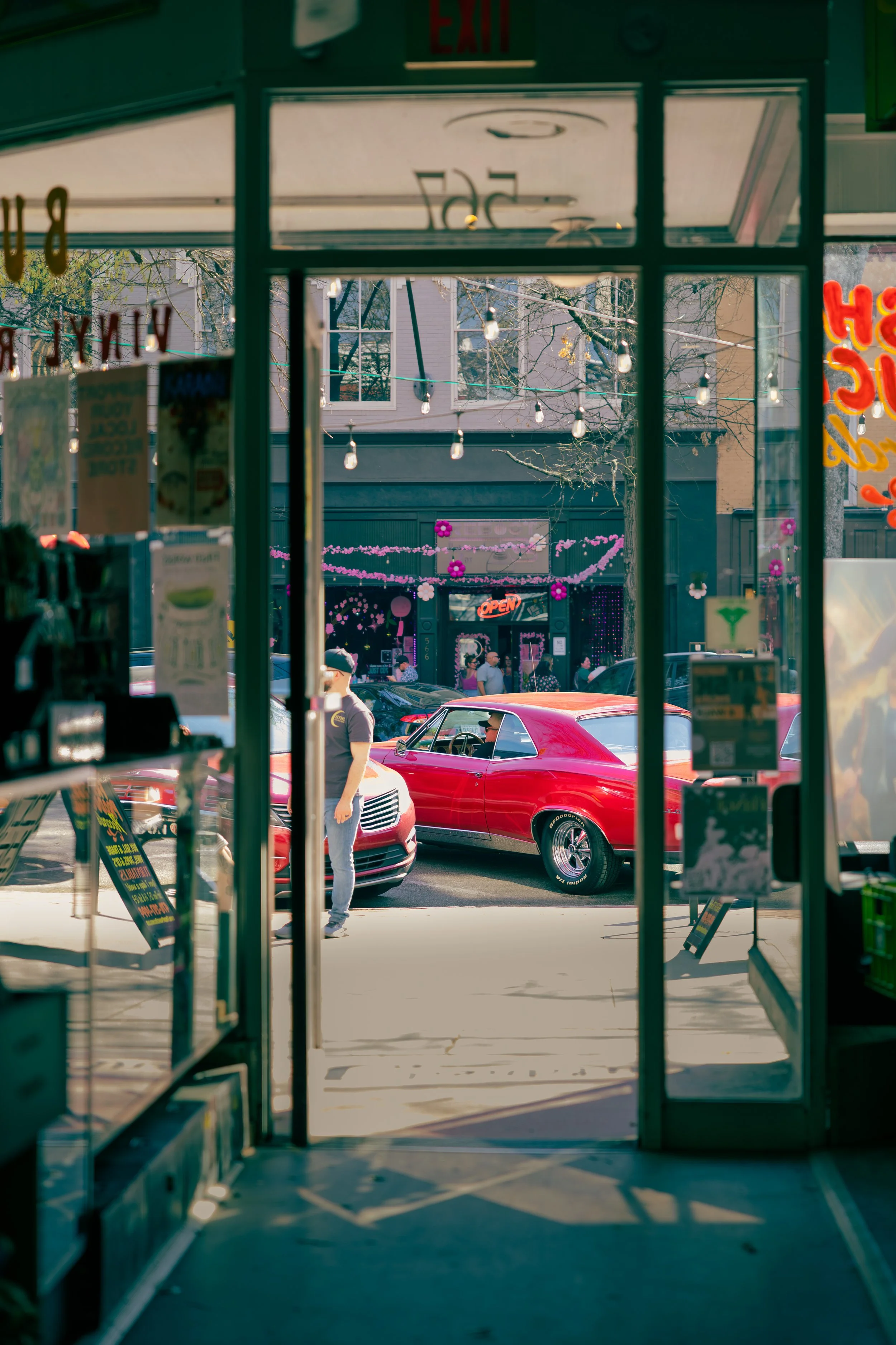 View through the entrance of a store looking out onto a street with a red classic car parked outside. The store interior has various posters and items, with a glimpse of a person in line. The exterior scene includes decorated storefronts with an 'Ope