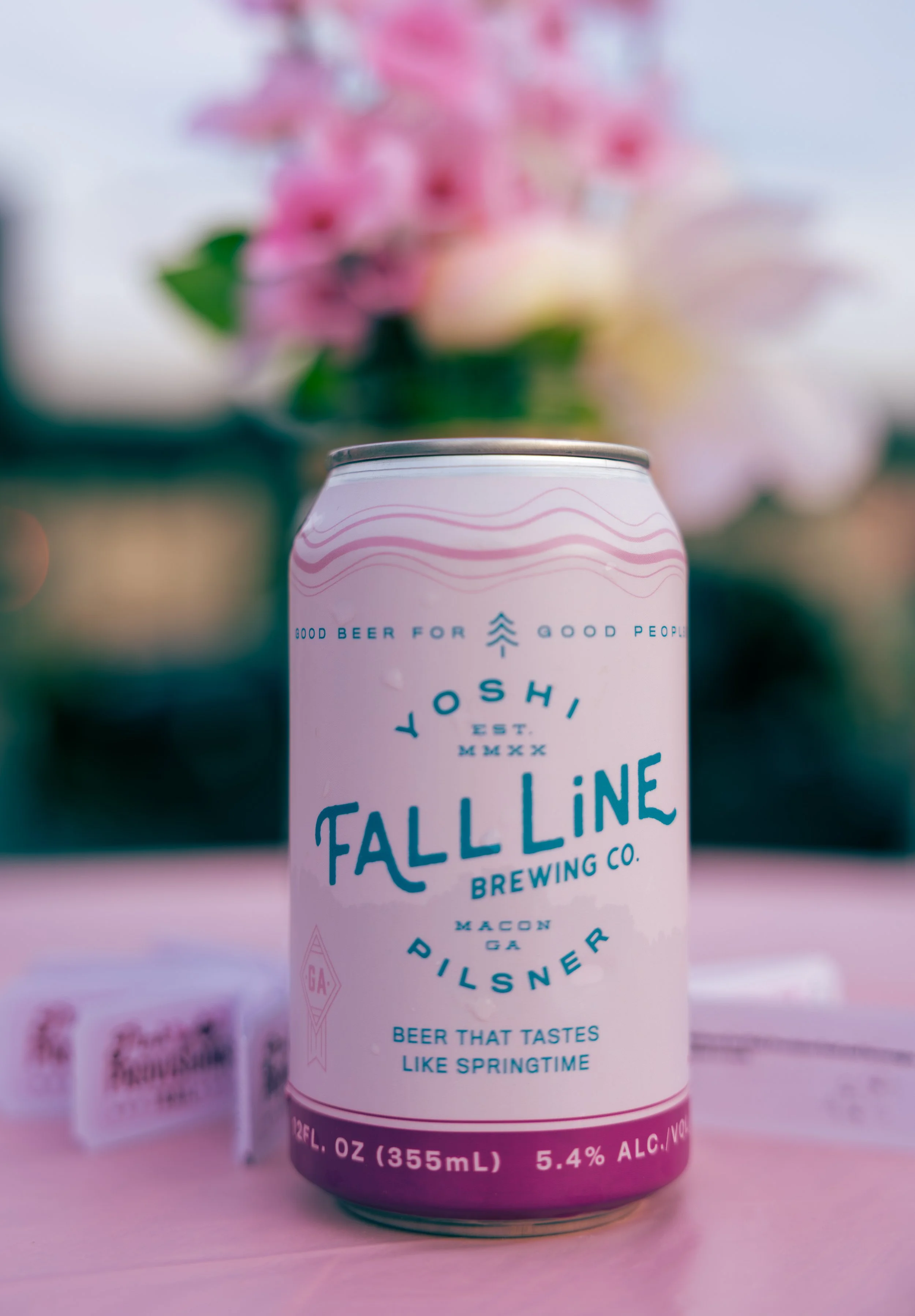 A can of Fall Line Brewing Co. Pilsner beer with a pink and white label, placed on a pink surface with blurred pink flowers in the background.