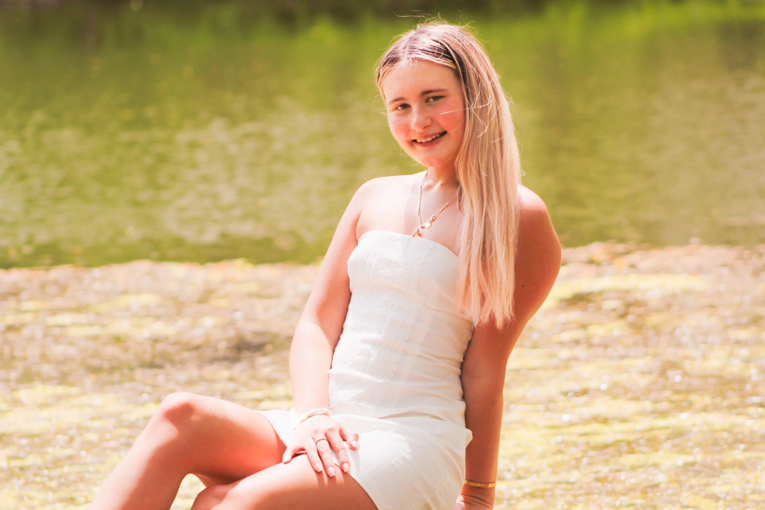 A girl with long blonde hair sitting by a body of water, smiling, wearing a white dress and jewelry.
