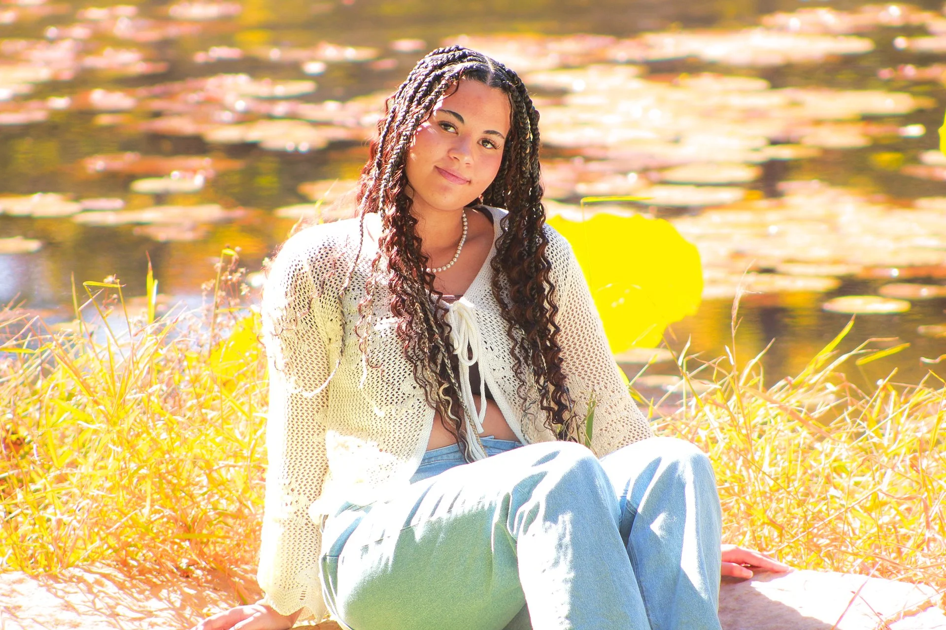 Young woman with long, curly hair sitting outdoors near water with lily pads, wearing a white crochet sweater, a black top, and light blue jeans, smiling gently.