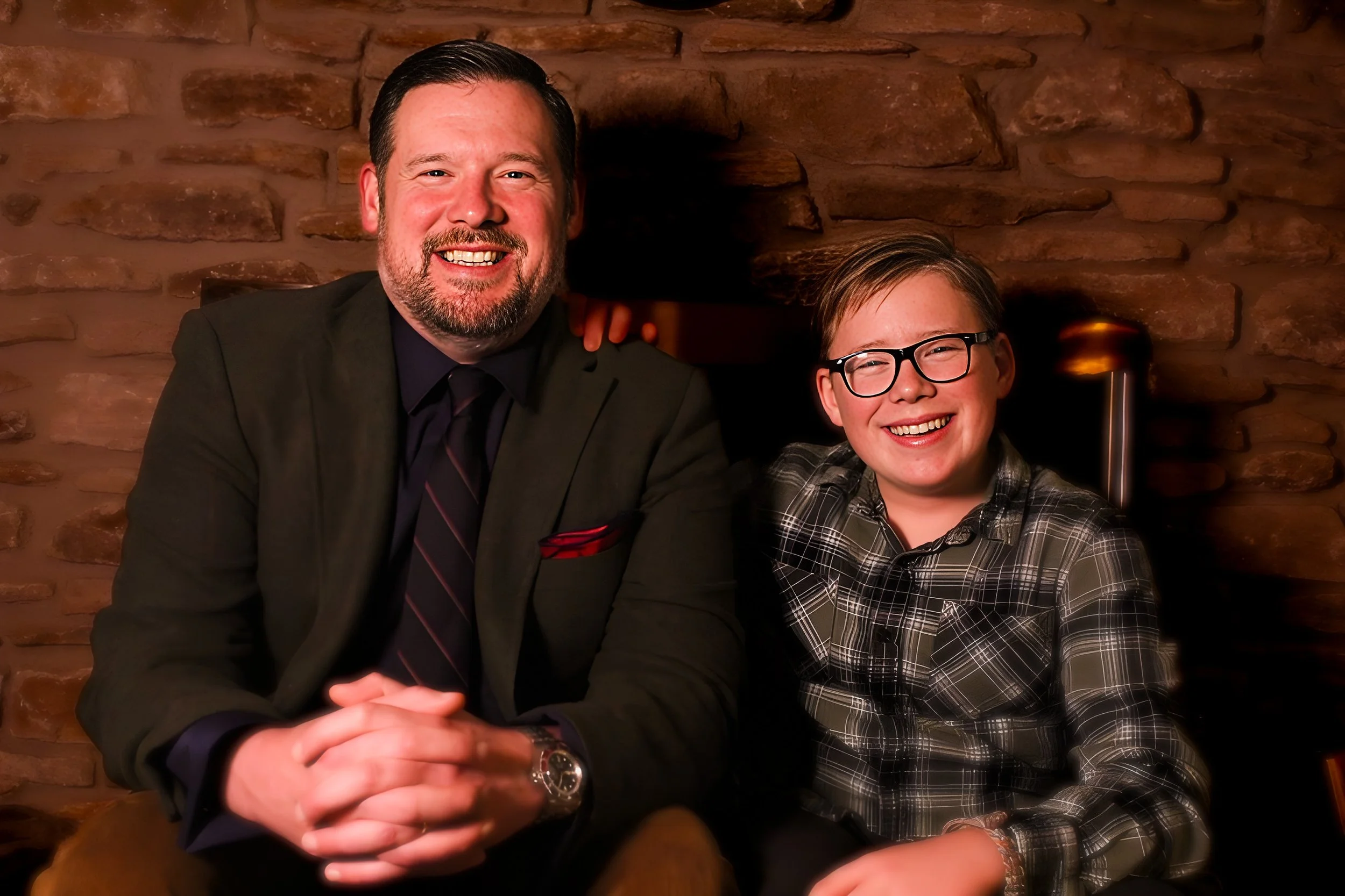 A man and a boy sitting together and smiling; the man has a beard and is wearing a dark suit with a tie, the boy has glasses and is wearing a plaid shirt; they are in a room with a brick wall background.