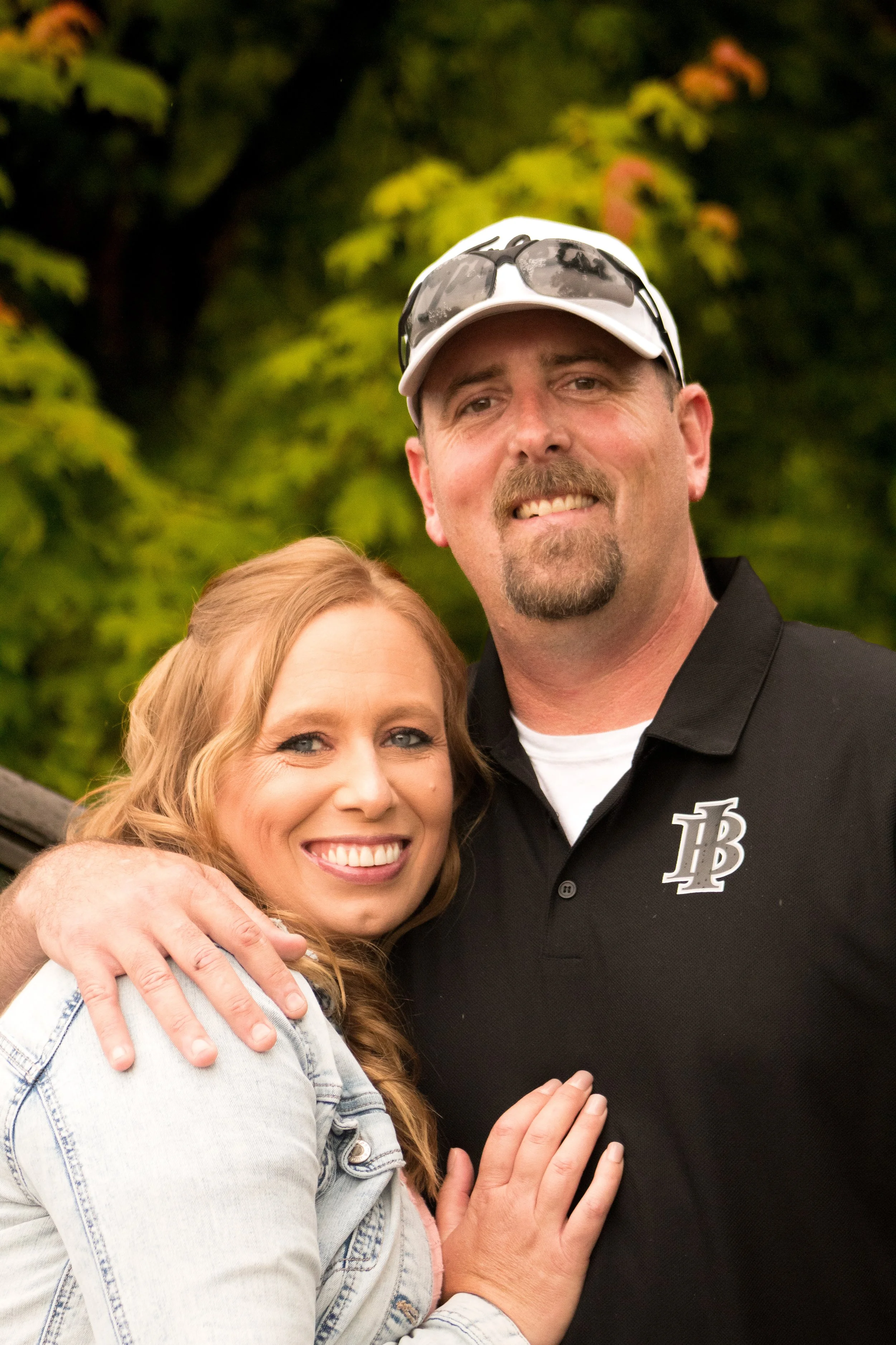 A smiling woman with light skin and blonde hair, wearing a denim jacket, hugging a man with light skin, a short beard, sunglasses on a white cap, and a black polo shirt with a logo, outdoors with green trees in the background.