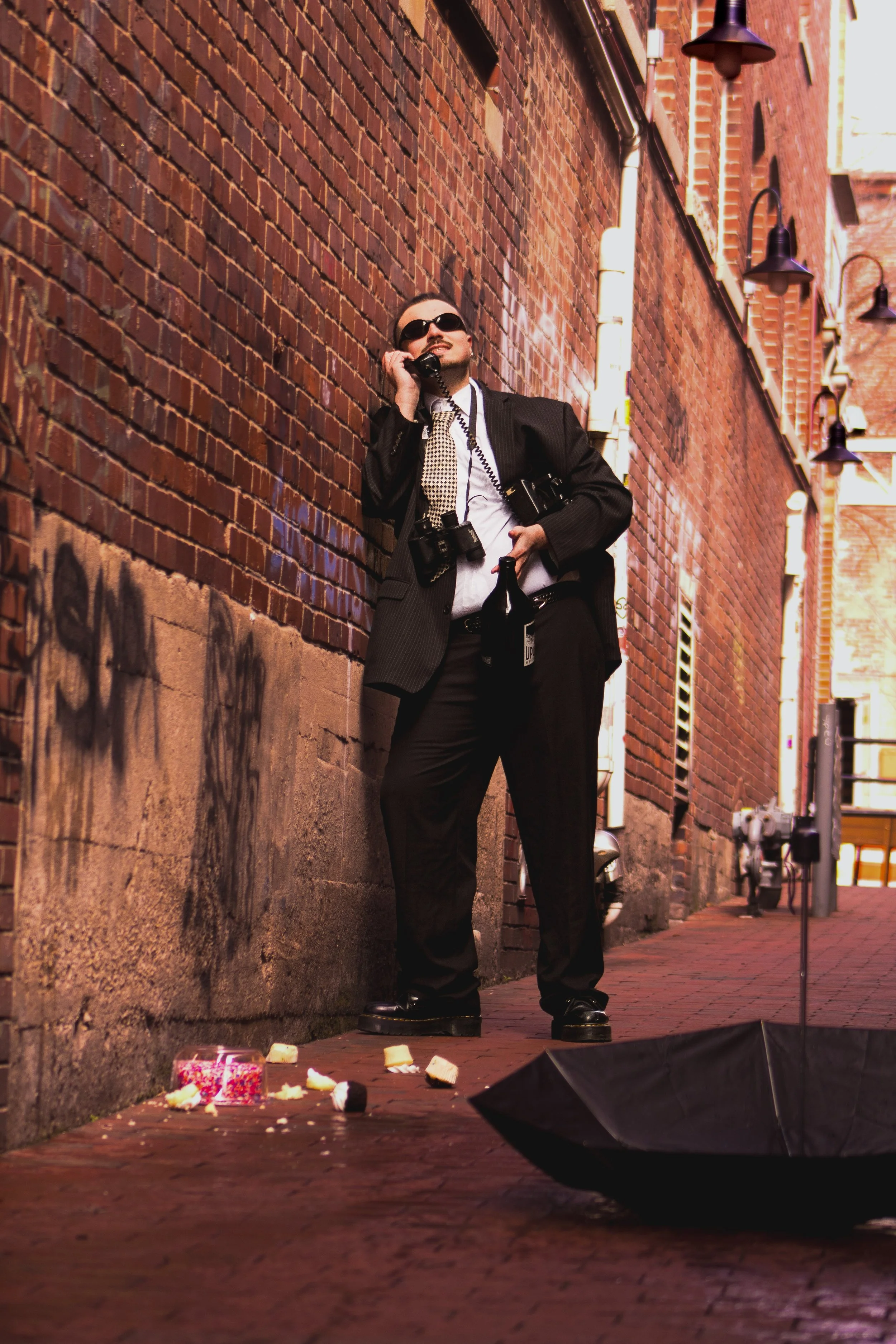 A man in a suit with sunglasses, a camera hanging around his neck, and a tie, standing against a brick wall in an alley, talking on a rotary phone while holding a bottle. There are birthday cake pieces and an overturned umbrella on the ground nearby.