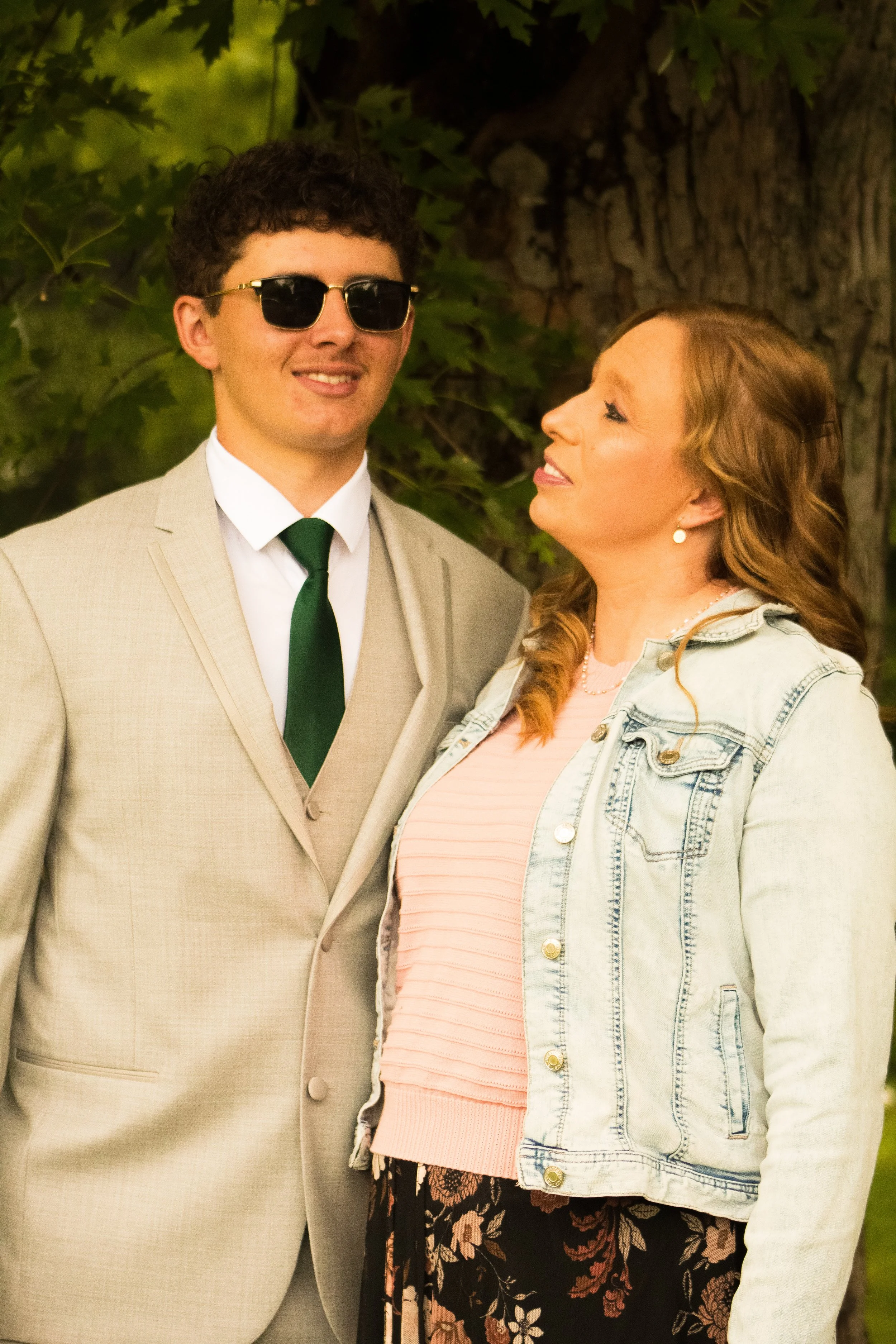 A young man wearing a beige suit, white shirt, green tie, and black sunglasses, standing next to a woman with long, wavy reddish-blonde hair wearing a denim jacket over a pink top and floral skirt, outdoors with a large tree and green foliage in the 