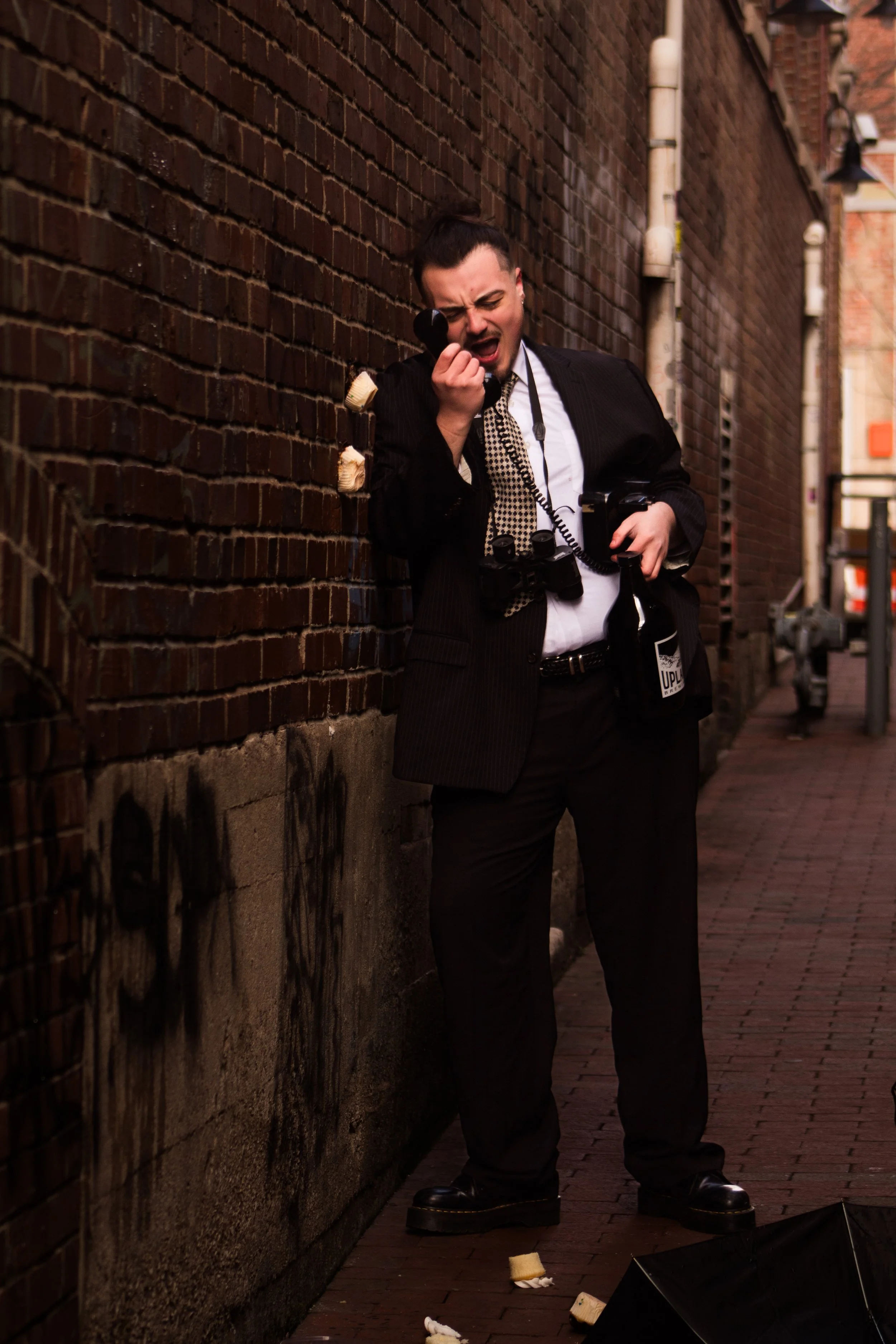 A man in a suit leaning against a brick wall, holding a phone to his ear with a distressed expression, surrounded by broken food pieces and an umbrella on the ground.