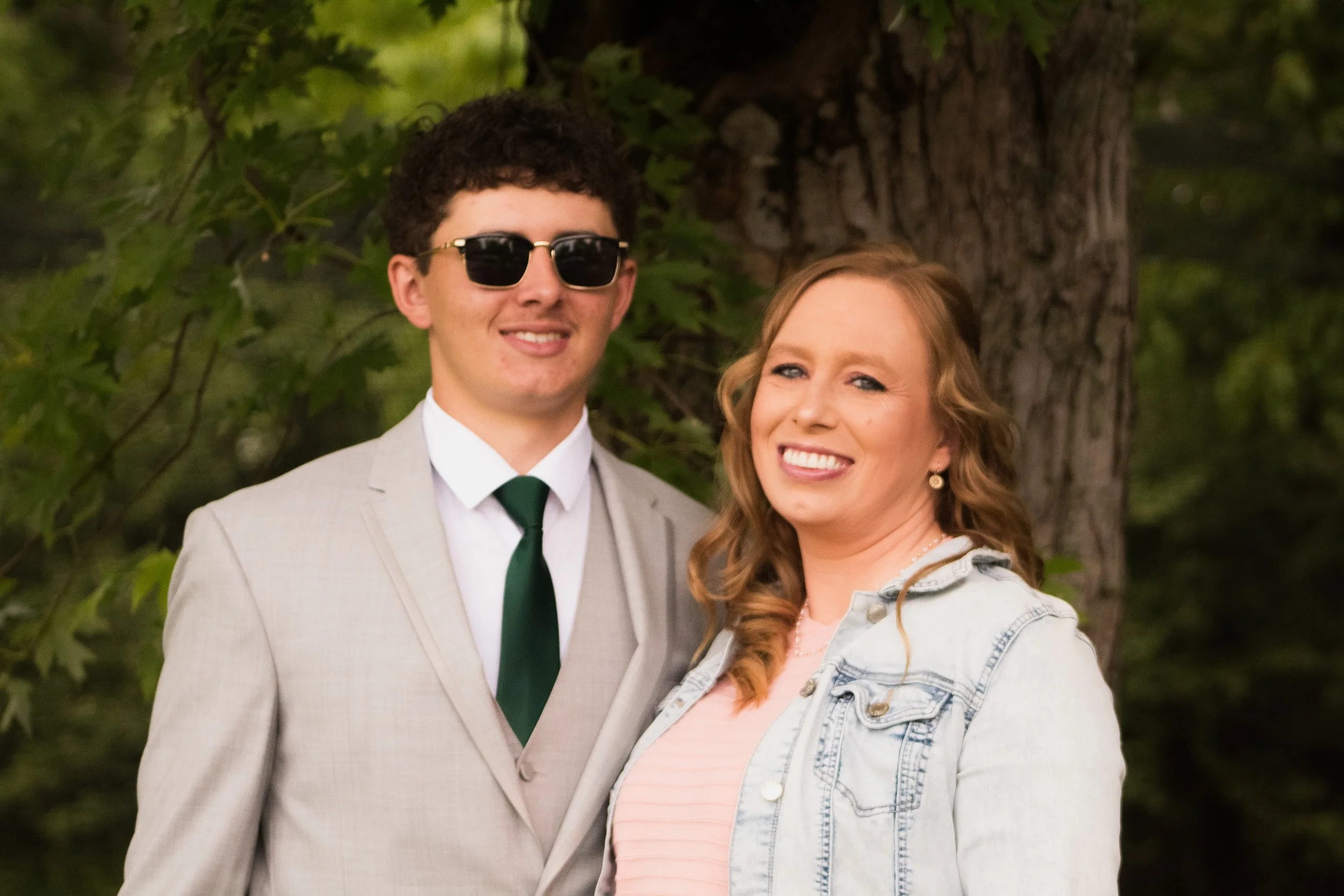 A young man in a light-colored suit with a dark green tie and sunglasses, standing next to a young woman with curly red hair wearing a light blue denim jacket and a pink striped shirt, smiling outdoors near a tree.