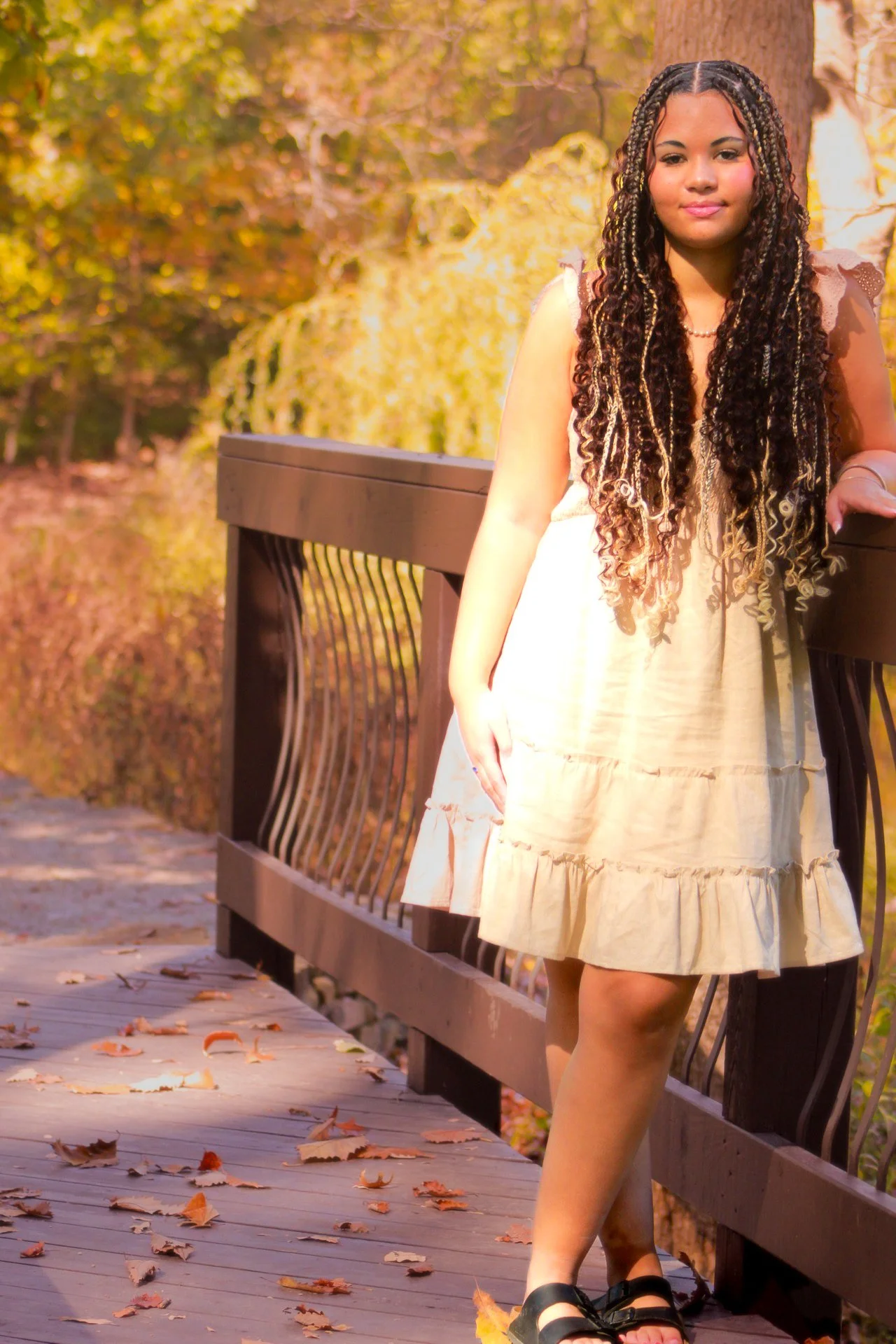 A young woman with long, curly hair leaning on a wooden railing on a bridge in a park during autumn.