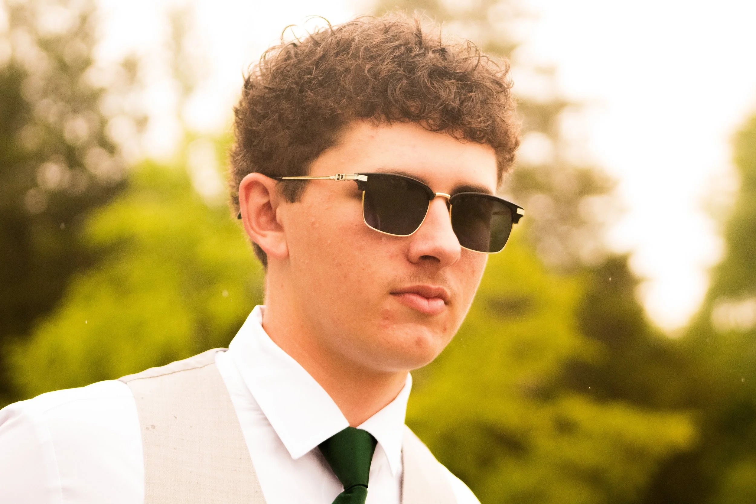 A young man with curly brown hair wearing black sunglasses, a white shirt, a beige vest, and a dark green tie, outdoors with blurred trees in the background.