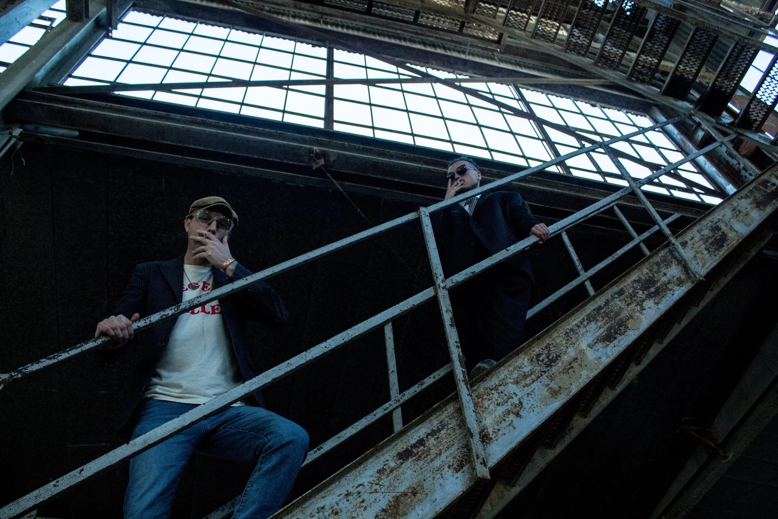 Two men standing on a rusty fire escape in an urban setting, one wearing sunglasses and a suit, the other wearing glasses and a casual jacket, both appear to be posing or looking at the camera.