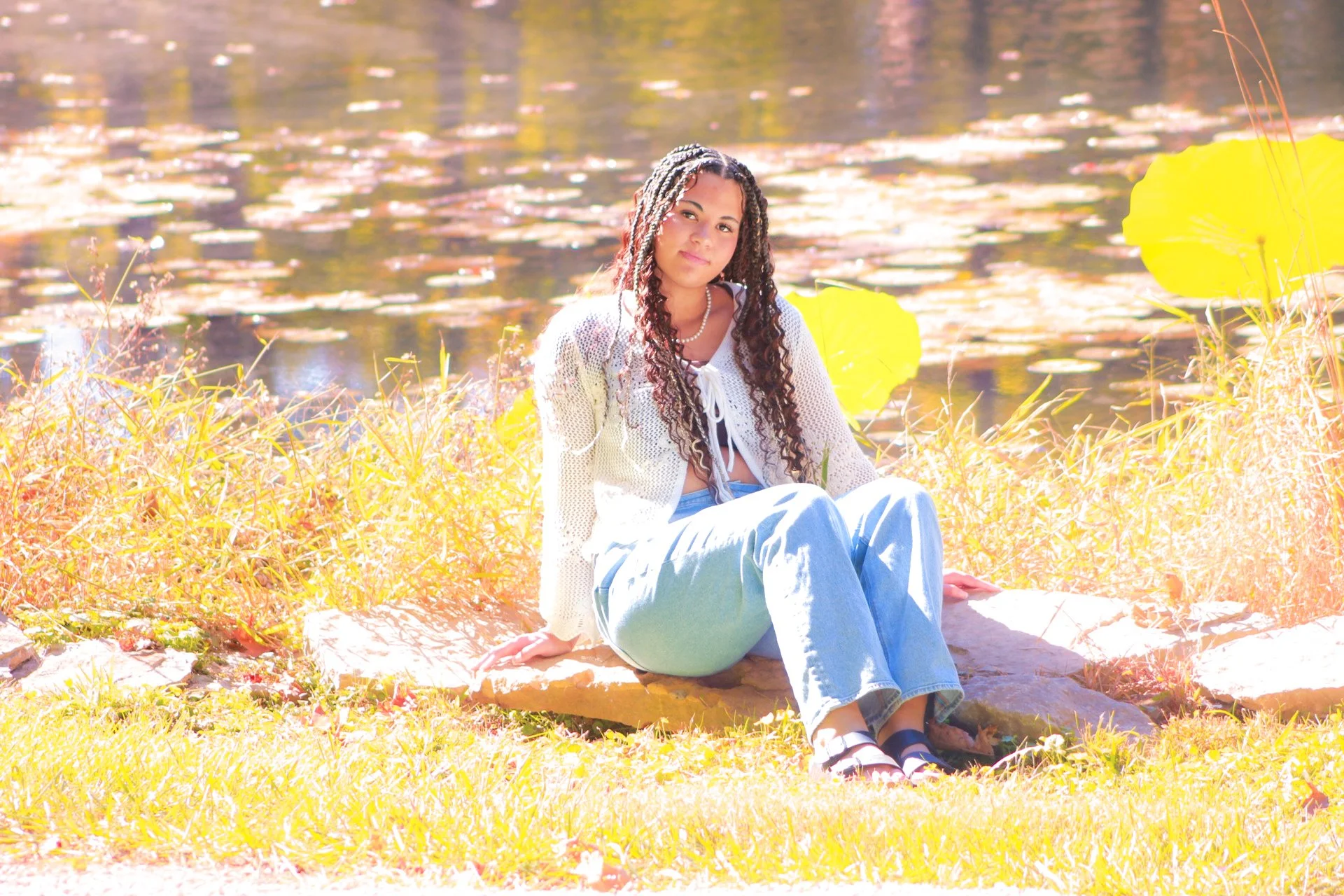 Young woman sitting on a rock near a pond with lily pads, surrounded by yellow foliage, wearing a white cardigan, light blue jeans, and black sandals.