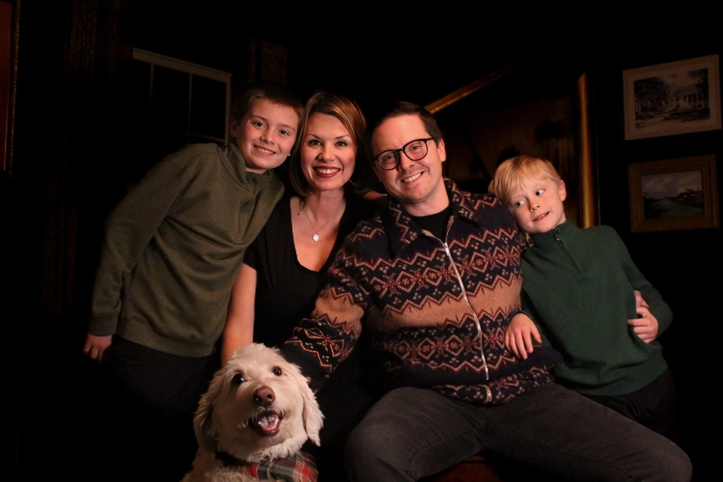 A family of five with two children and a dog posing indoors in a dimly lit room, smiling at the camera.