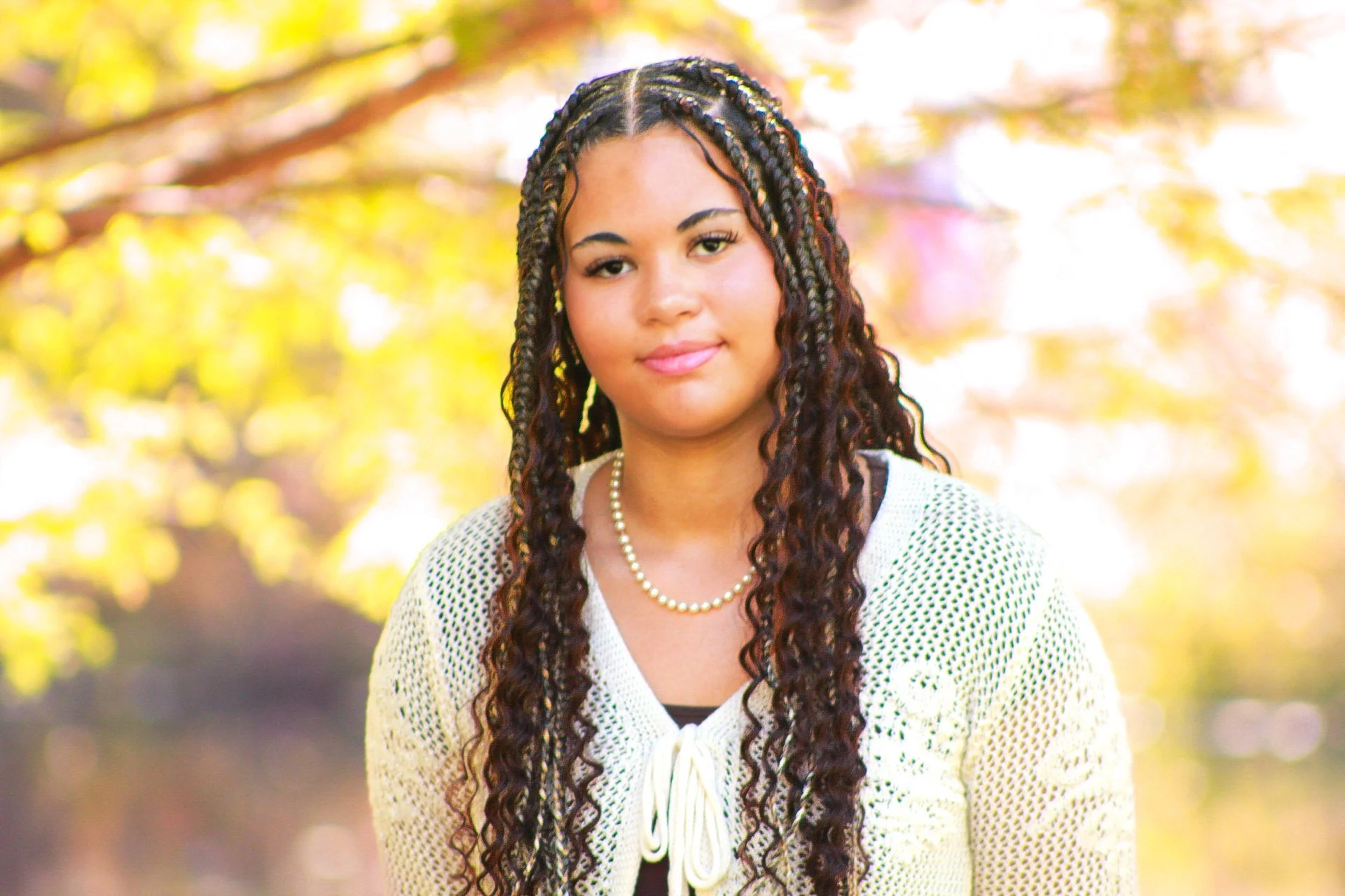 A woman with long, dark, curly braids standing outdoors with blurred autumn leaves in the background.