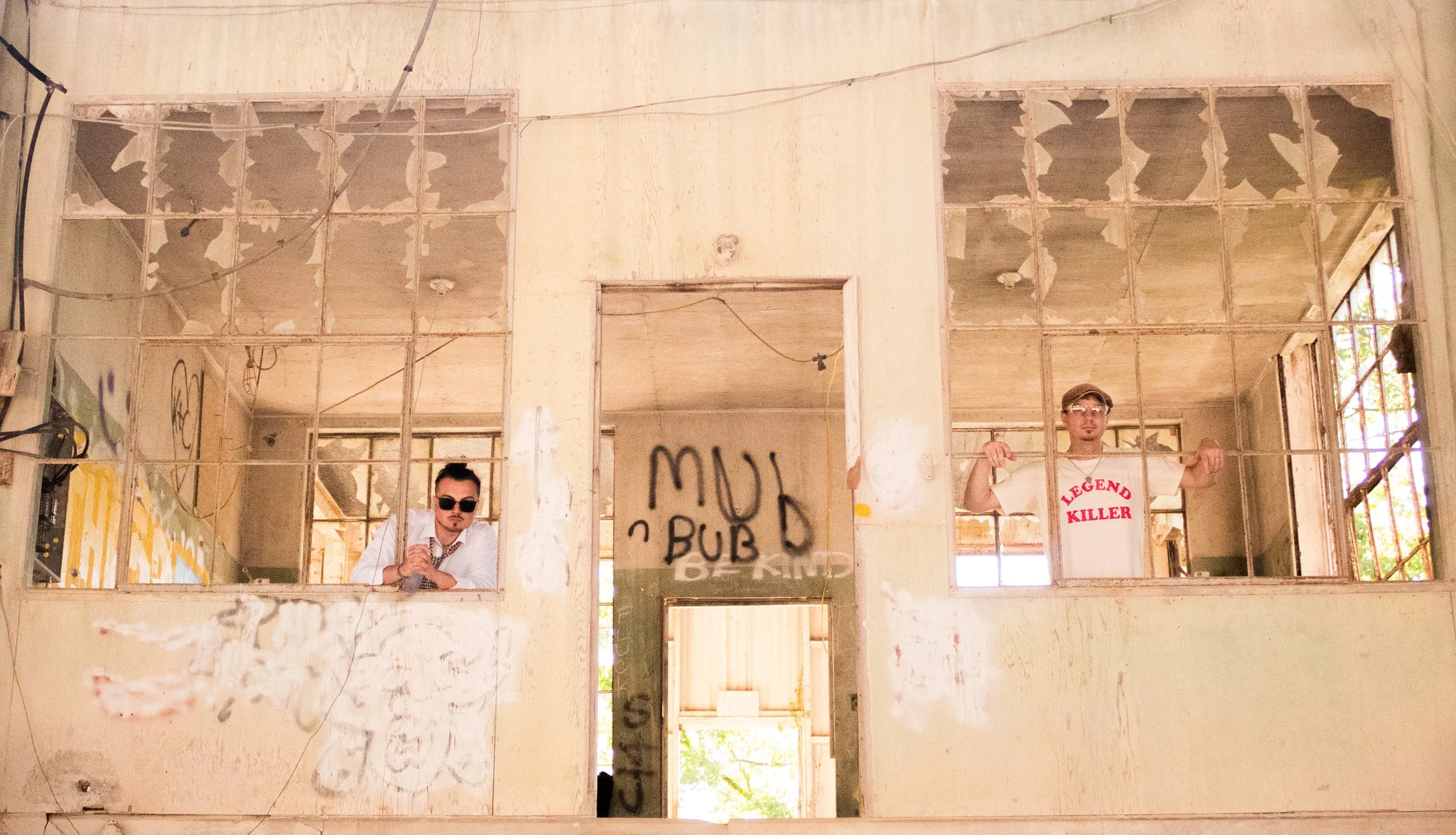 Two men in an abandoned building with peeling walls and graffiti, standing behind large geometric window frames. One man wears sunglasses and a white shirt; the other wears glasses and a white T-shirt with red text.
