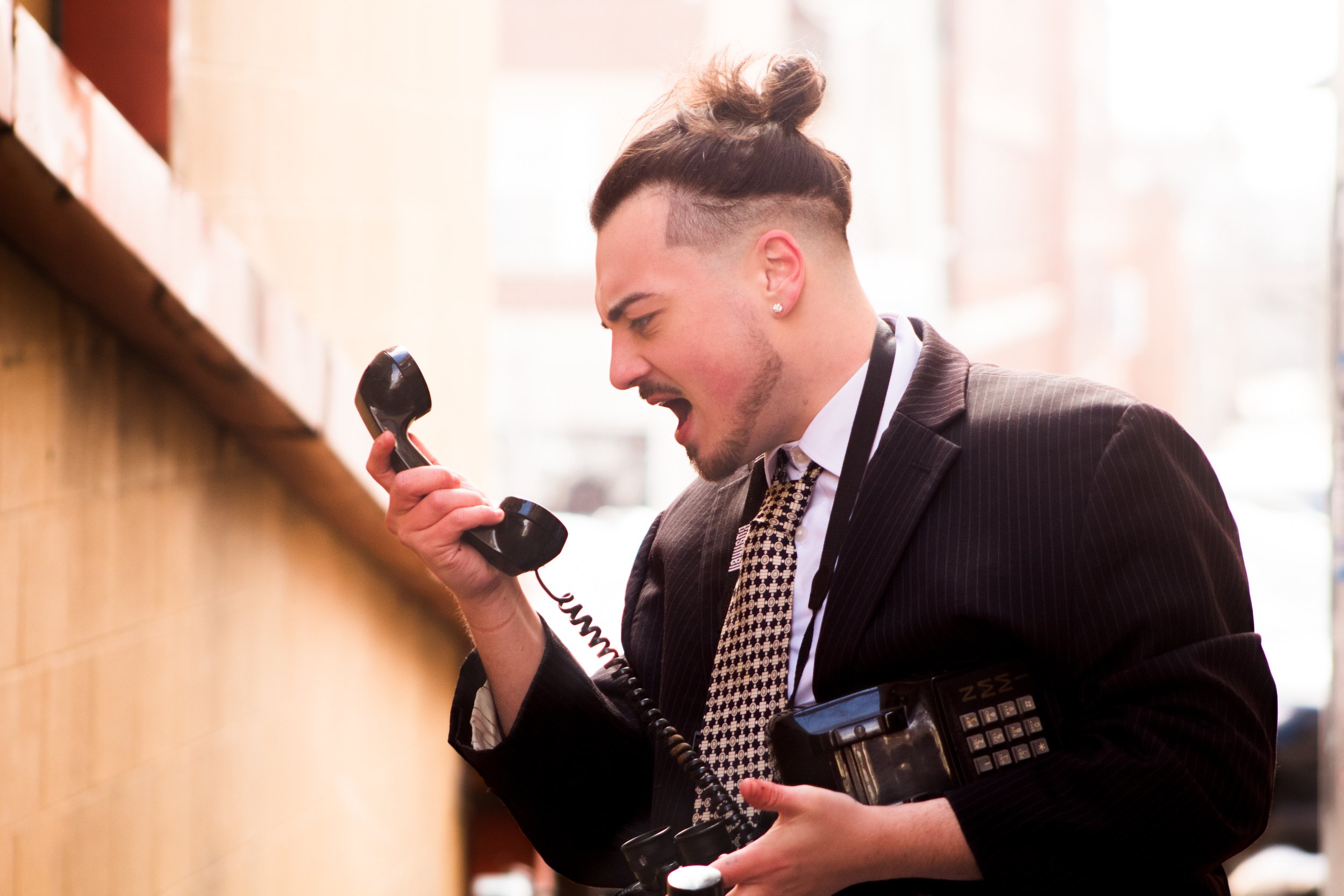 A man in a suit and tie yelling into a telephone with a distressed expression, holding a vintage camera in his left hand.