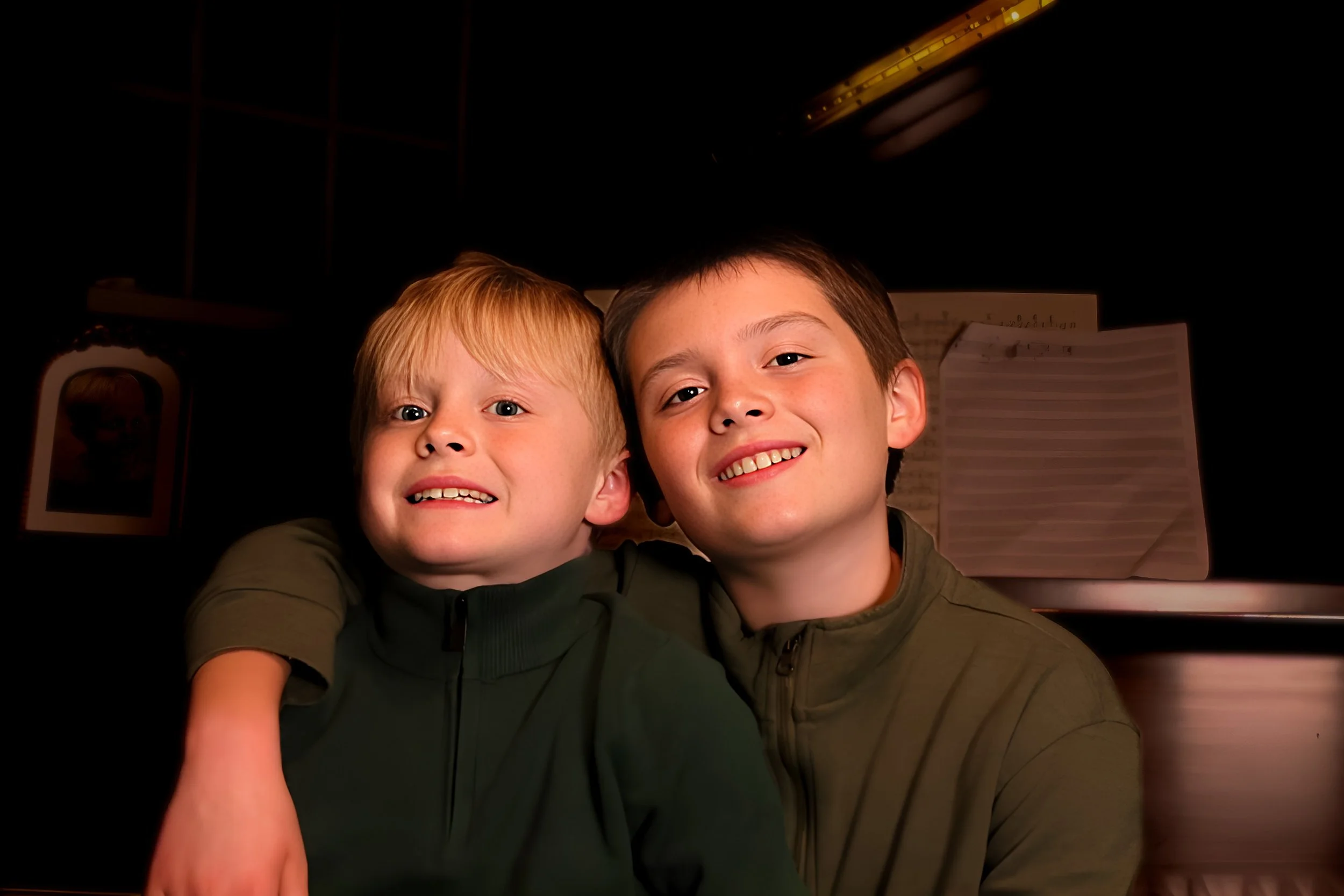 Two boys smiling and hugging each other in front of a dark background, with sheet music visible behind them.