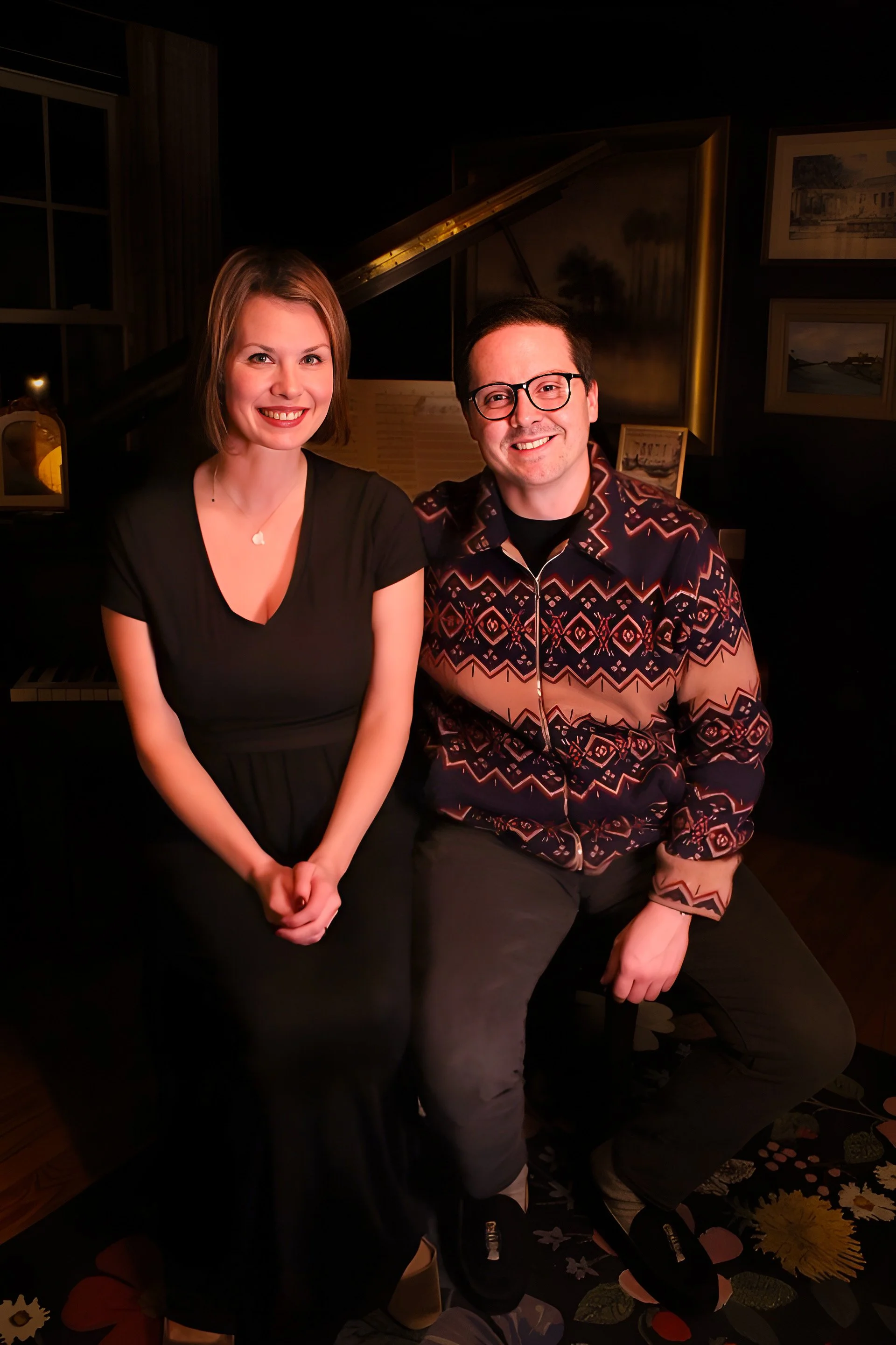 A young woman and a young man sitting together in a dimly lit room with a grand piano in the background. The woman has short, light brown hair and is wearing a black dress, smiling at the camera. The man has short dark hair, glasses, and is wearing a