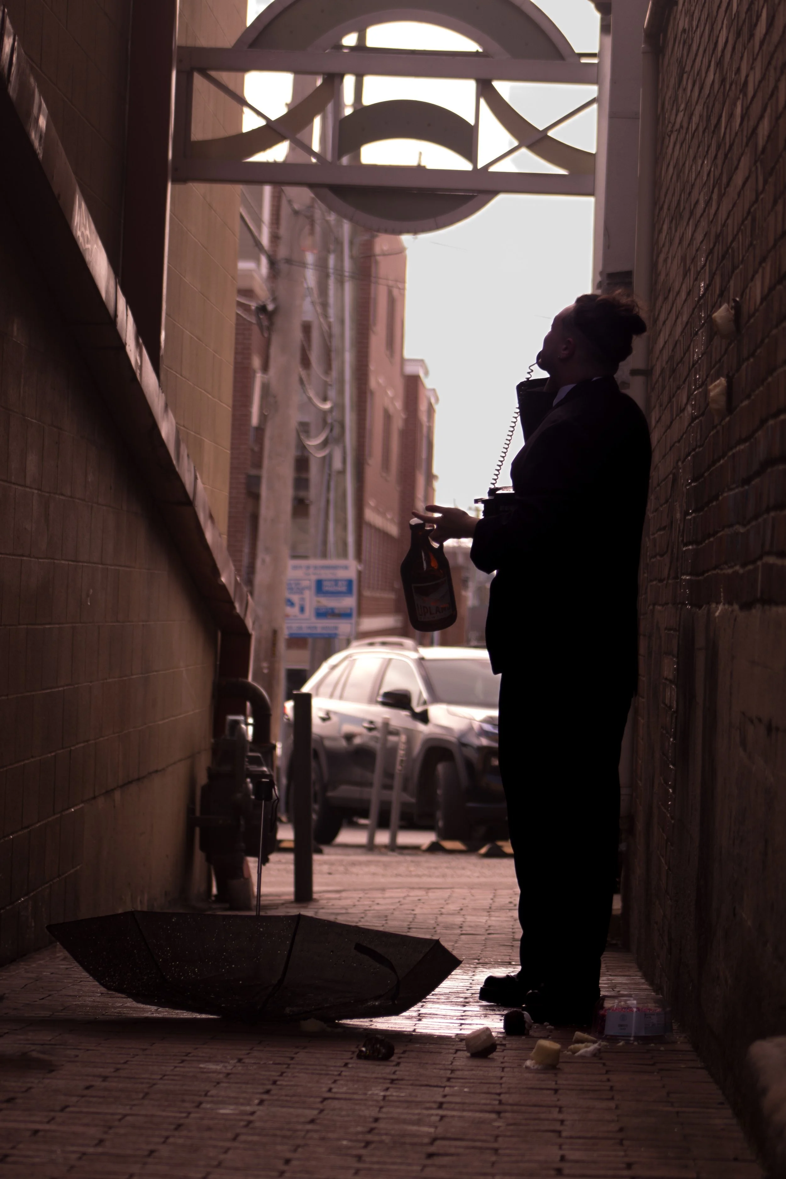 Silhouette of a person standing in a narrow alleyway holding a bottle and an umbrella lying on the ground, with a car and city buildings in the background.