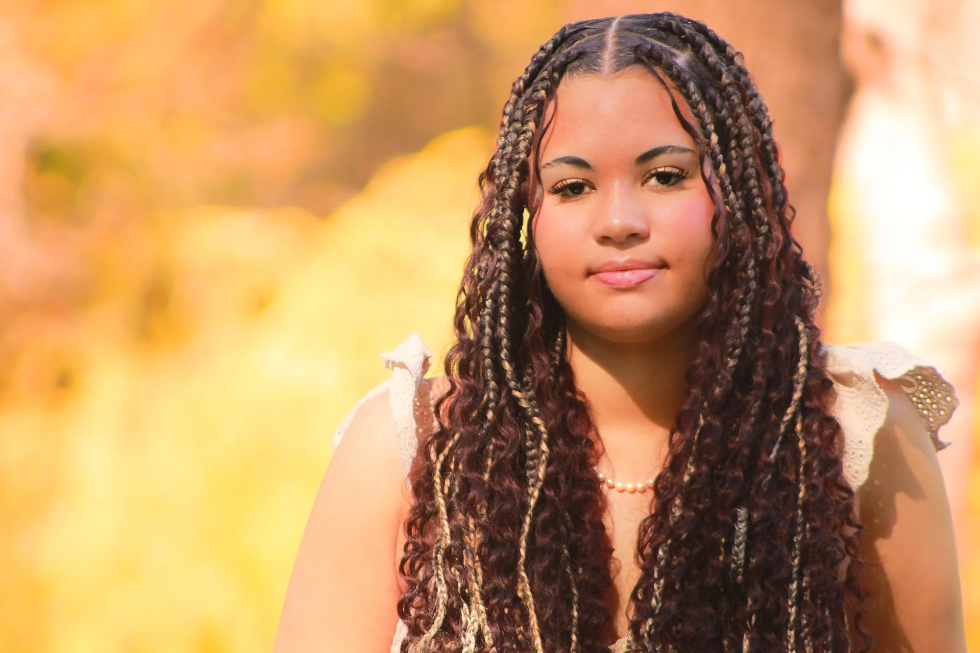 A young woman with long, dark, curly and braided hair. She is wearing a cream-colored top with lace details on the shoulders and a pearl necklace. She is looking at the camera with a slight smile, outdoors in autumn, with blurred yellow and orange le