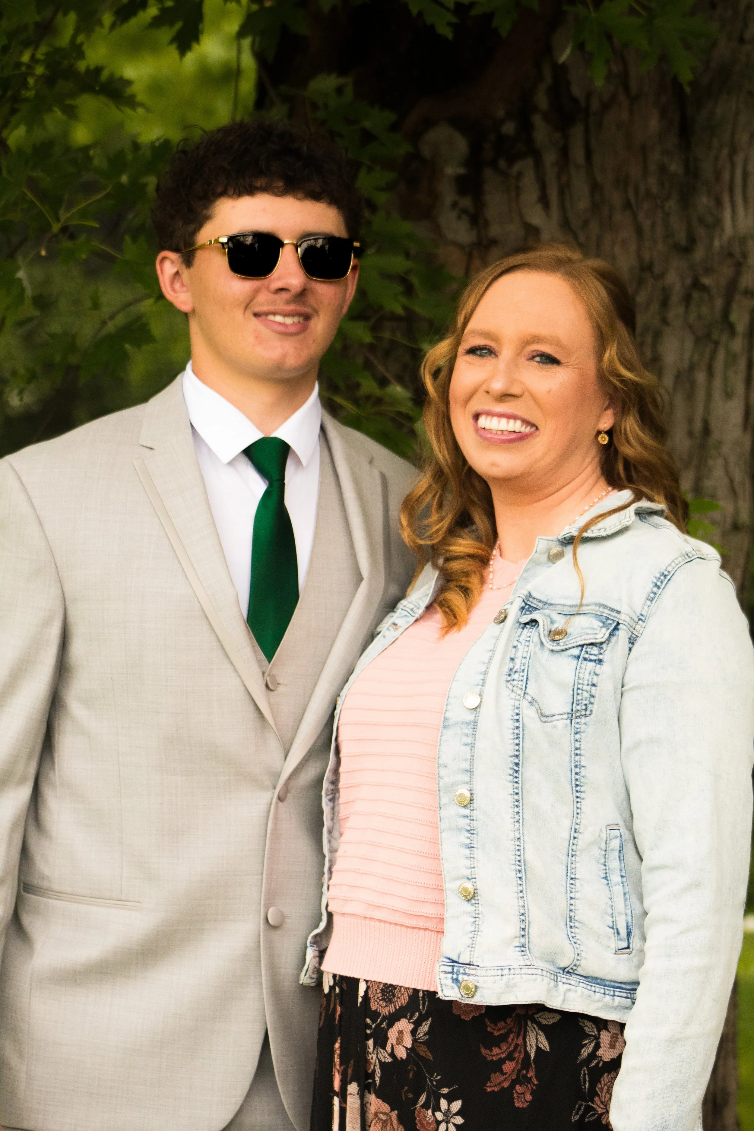 A man wearing a beige suit, sunglasses, a white shirt, and a green tie standing next to a smiling woman with wavy red hair, wearing a denim jacket and a pink top, standing outdoors near a tree with green leaves.