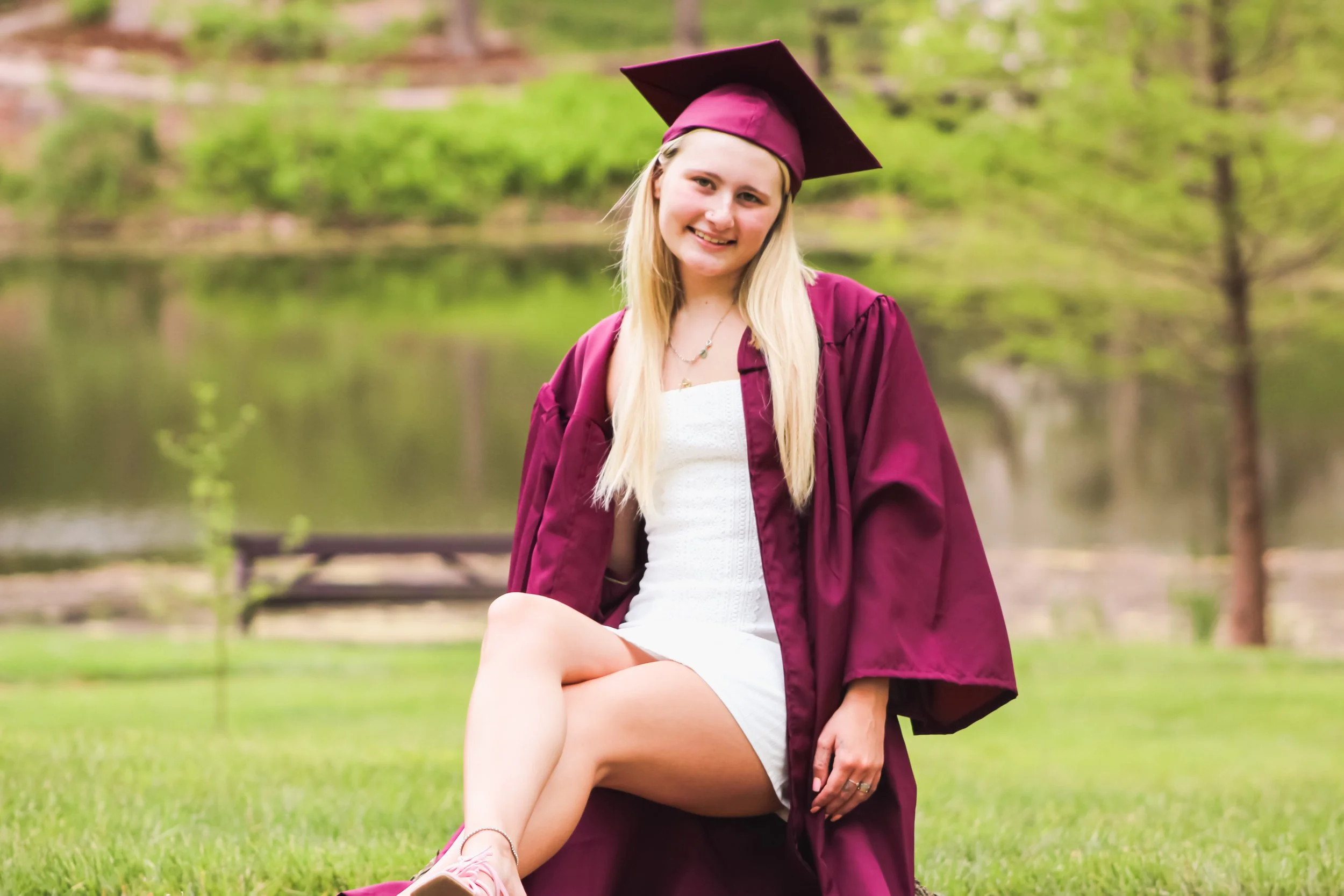 Young woman in graduation cap and gown sitting on grass near a body of water and trees, smiling at the camera.