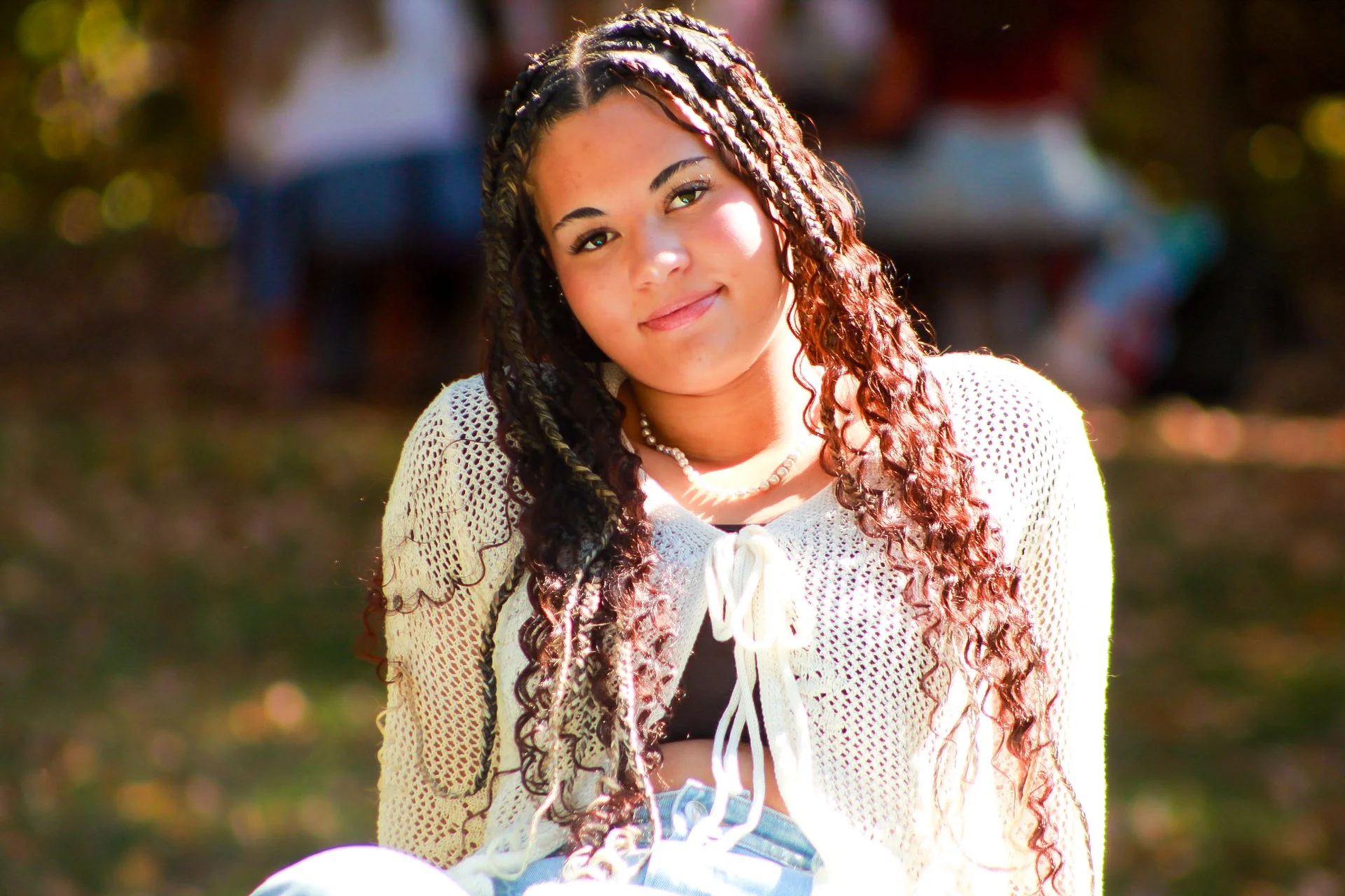 A young woman with long, curly hair and tan skin, wearing a white crochet cardigan and black top, posing outdoors with sunlight highlighting her face, and blurred colorful background.