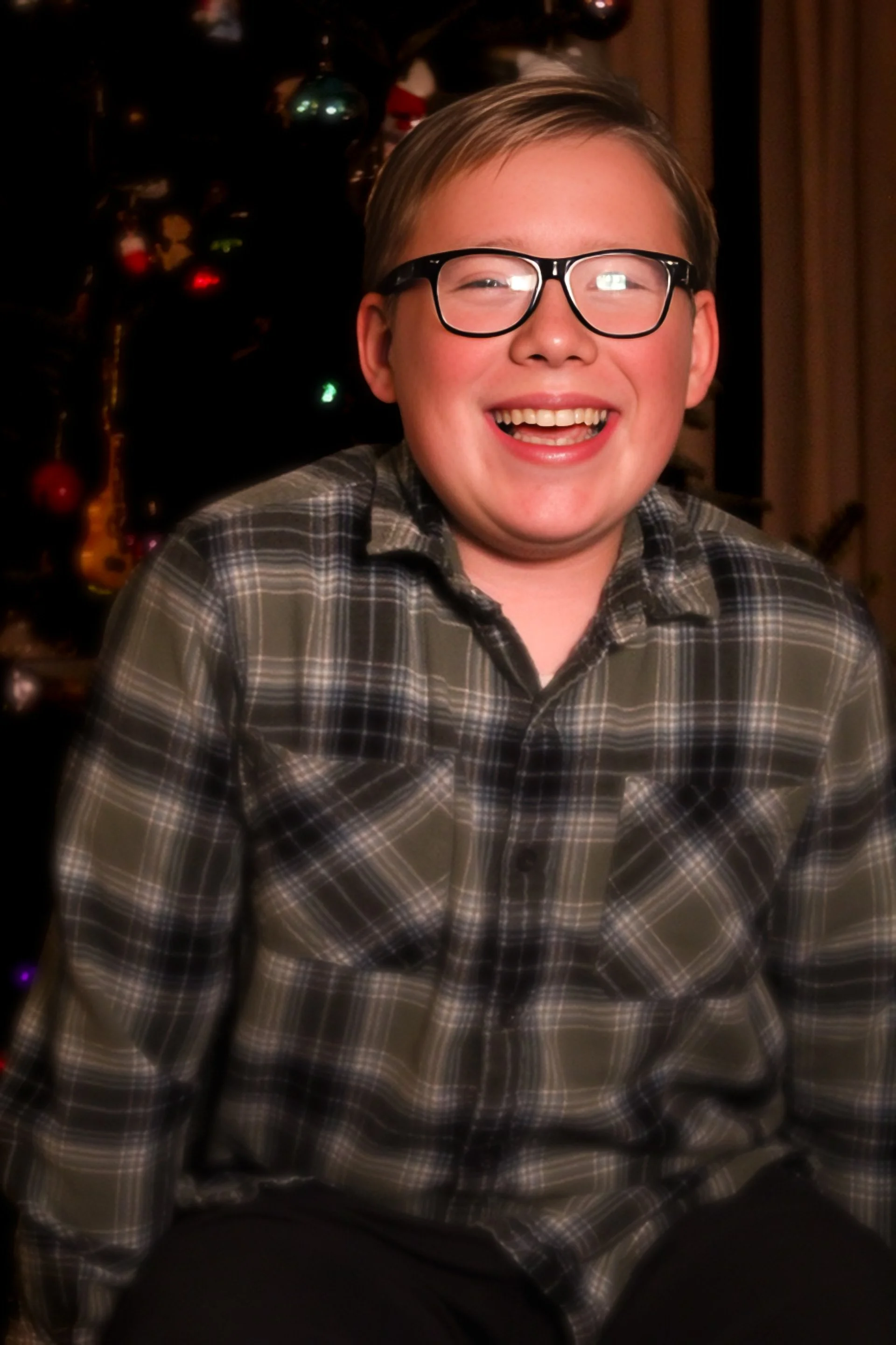 A young boy with glasses wearing a plaid shirt, smiling, sitting in front of a decorated Christmas tree.