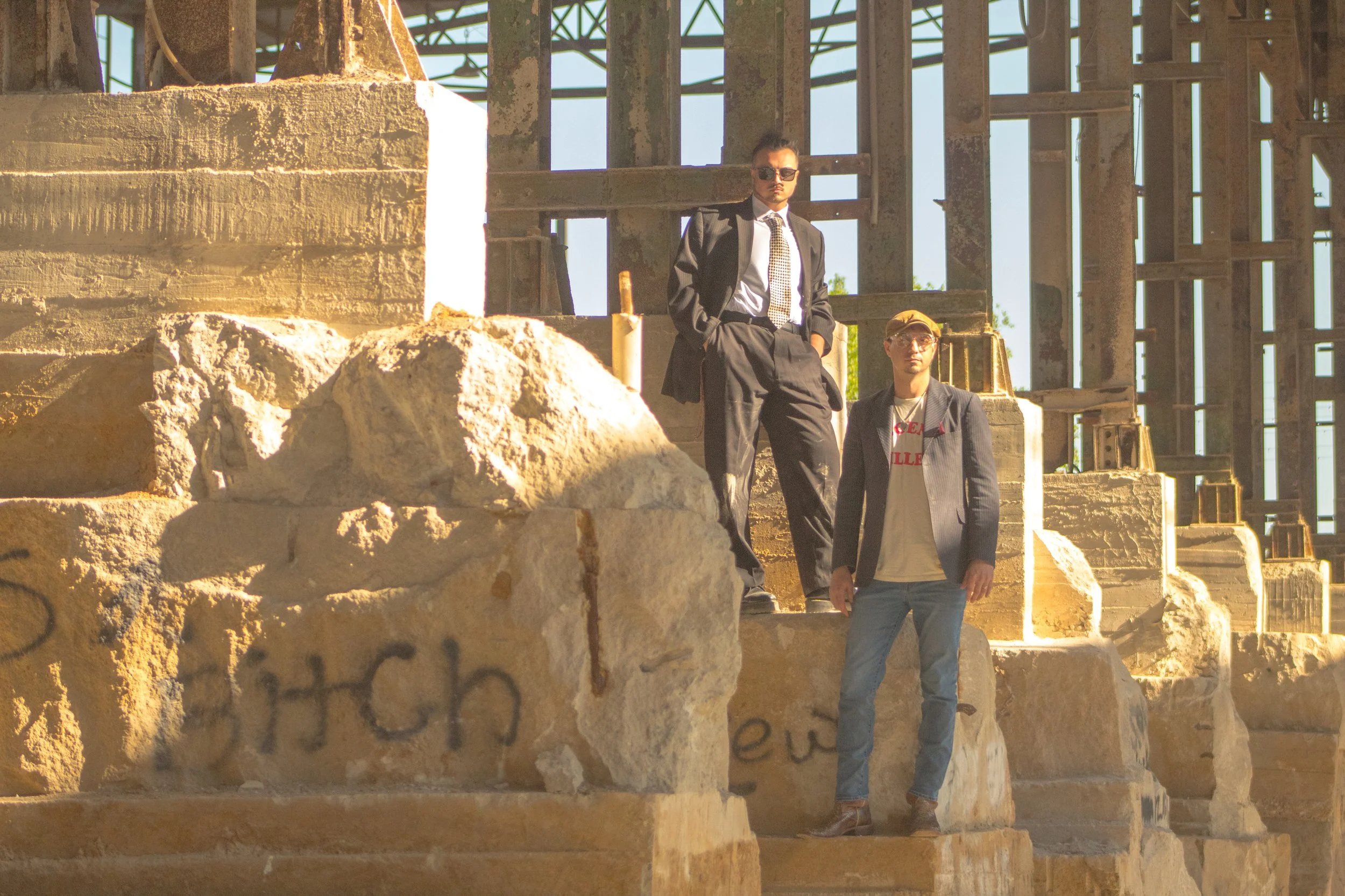 Two men standing on the ruins of a building under construction, with exposed concrete and metal framework in the background. One man is wearing a suit and sunglasses, and the other is wearing casual attire with a baseball cap.