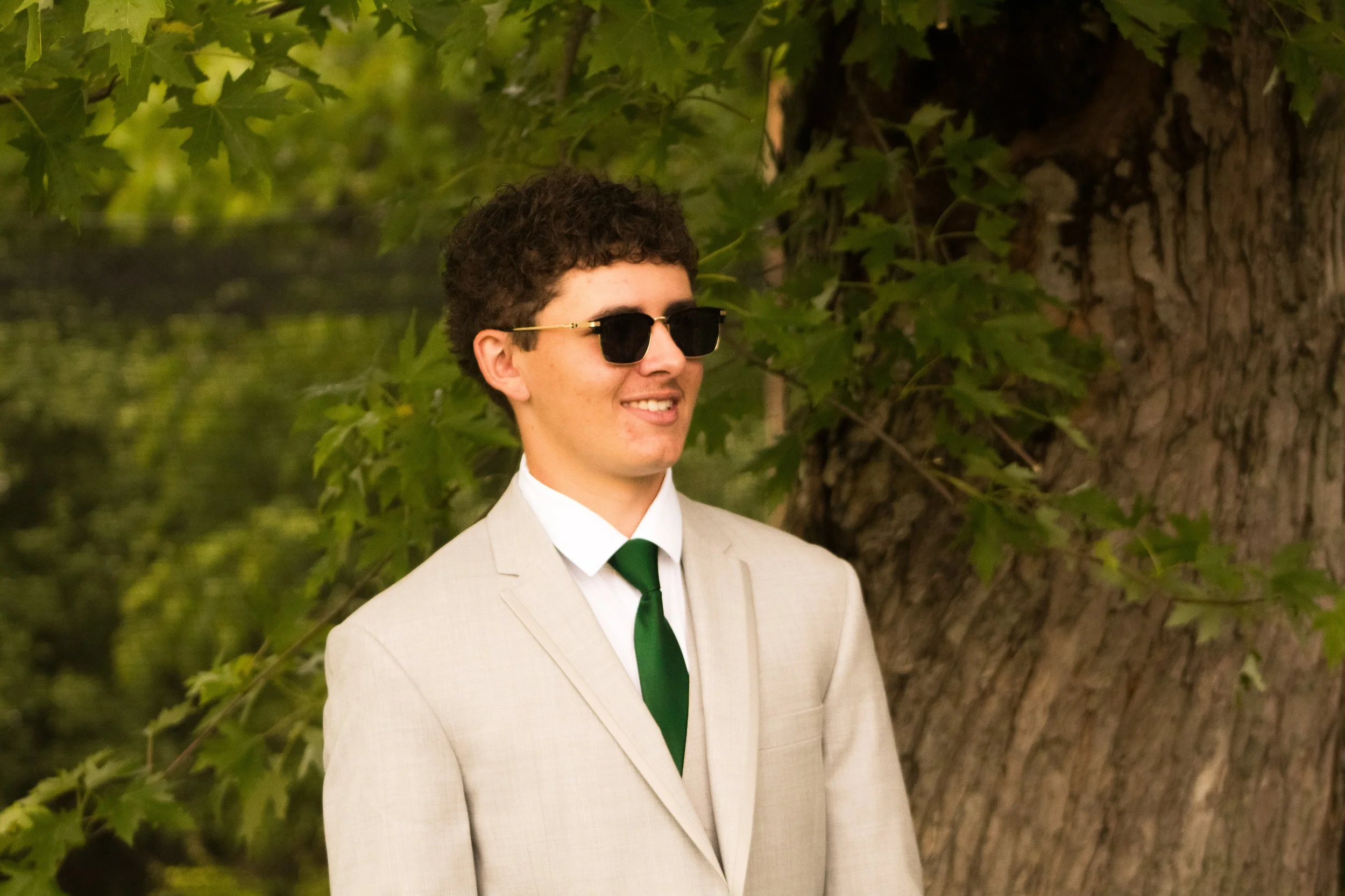A young man with curly hair and sunglasses wearing a beige suit with a white shirt and green tie, standing outdoors next to a large tree with green leaves in the background.