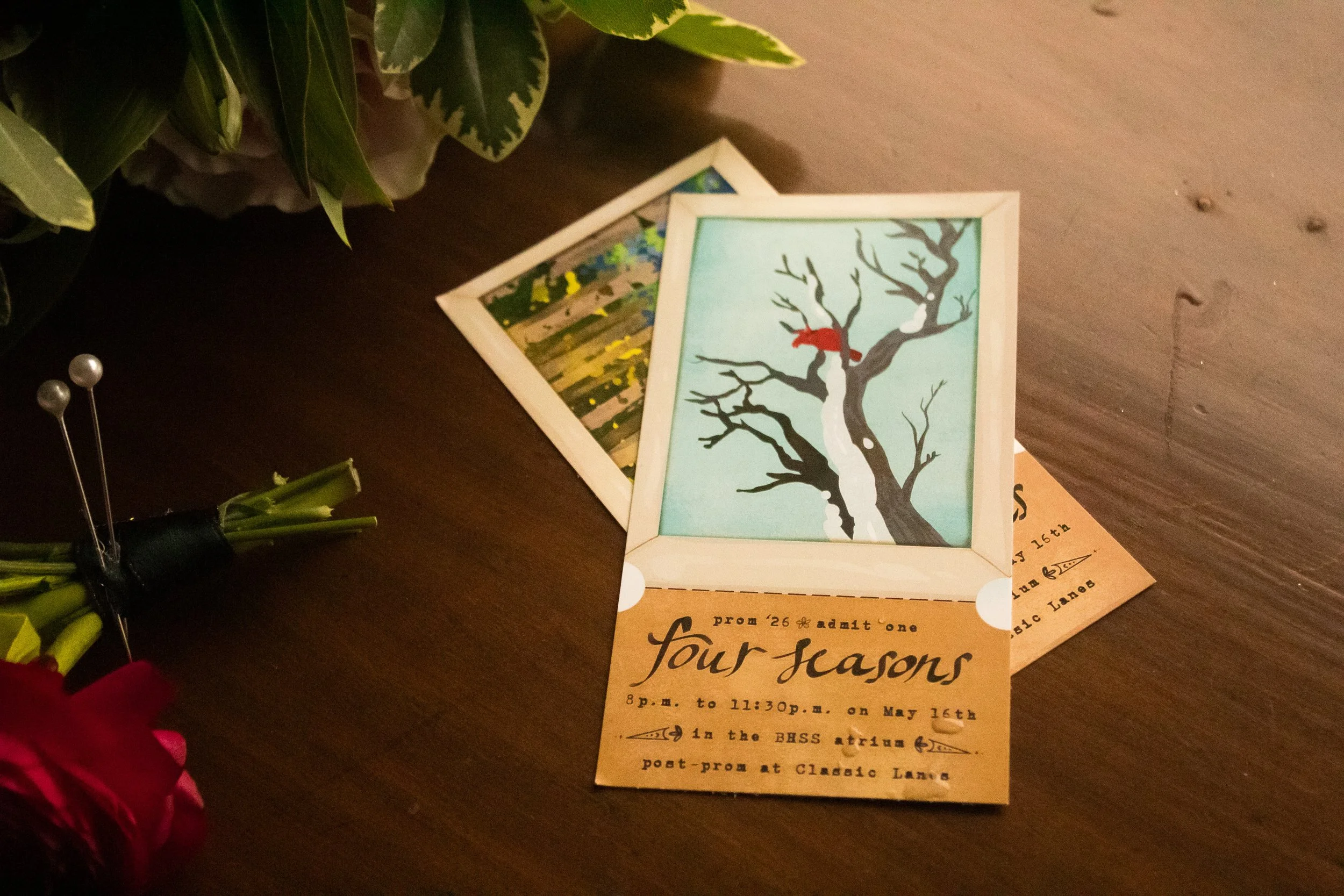 Two framed illustrations of winter trees, one with a red bird, are on a wooden surface next to a yellow event ticket for a prom, partially visible, with text mentioning prom dates and times. In the corner, part of a green leafy plant is visible.