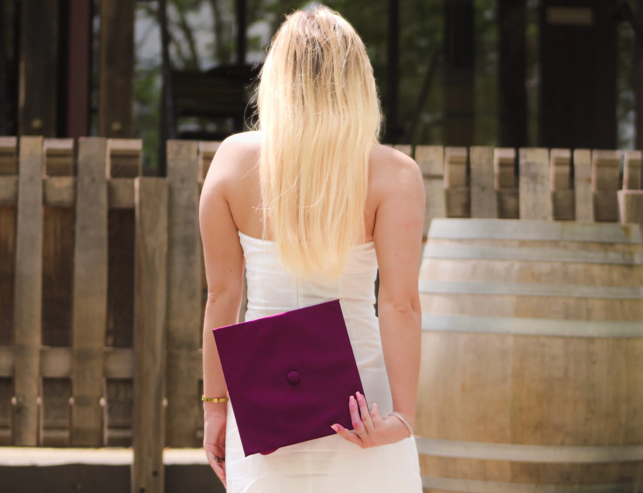 A woman with long blonde hair wearing a white dress and holding a purple folder behind her back, standing outdoors near a wooden fence and a hot tub.