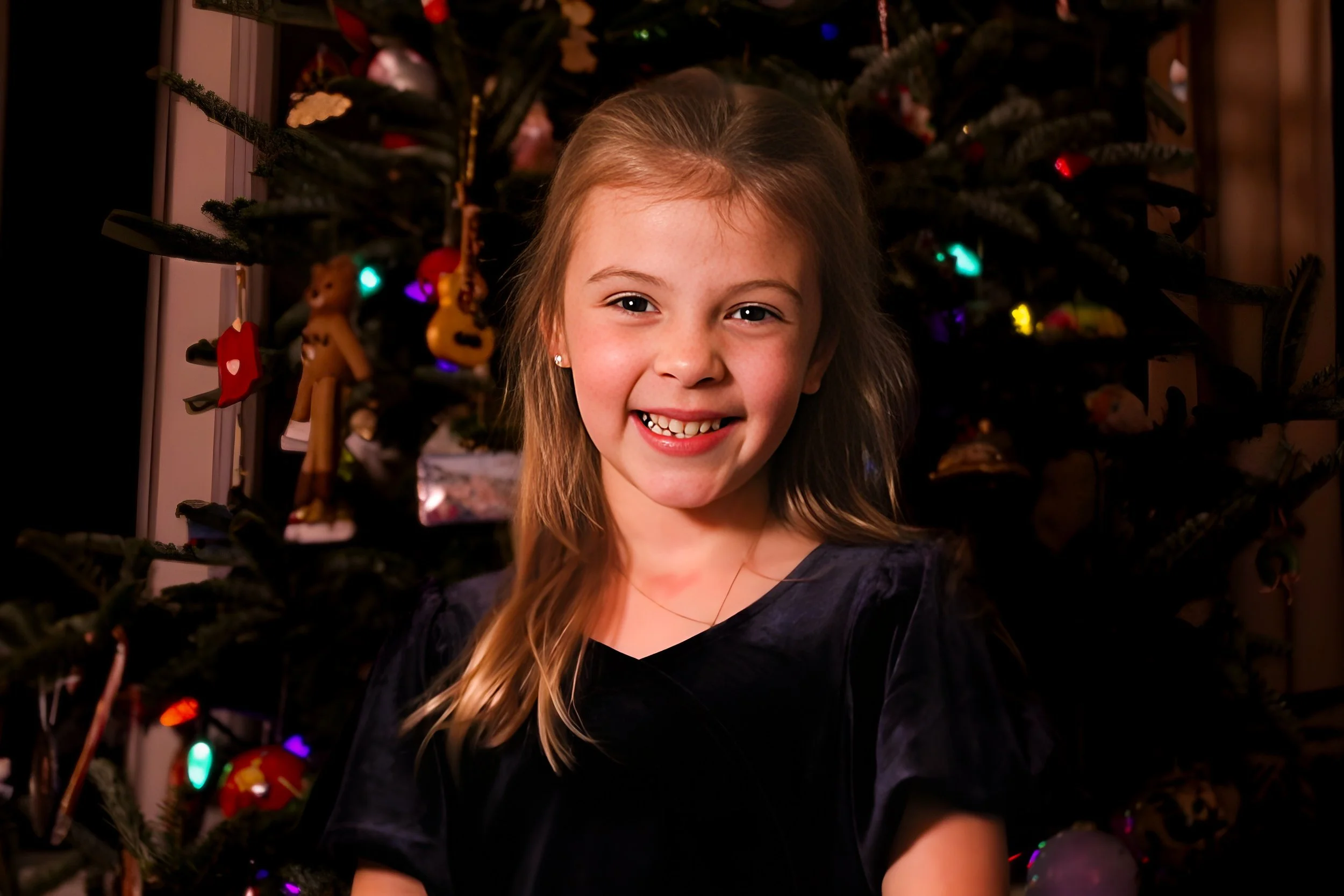 A smiling young girl with long blonde hair, wearing a dark dress, standing in front of a decorated Christmas tree with colorful ornaments and lights.