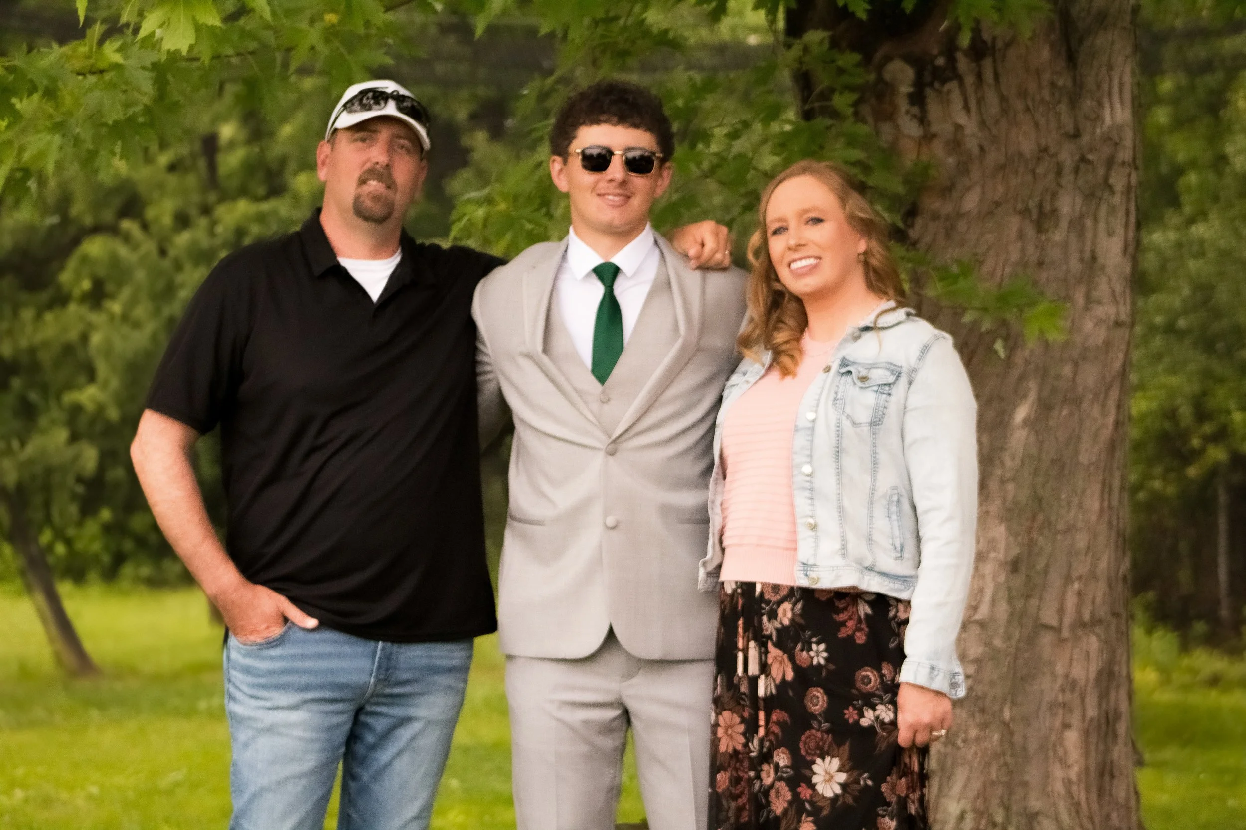 Three people standing outdoors in front of a large tree with green leaves. The person on the left is a man wearing a black shirt, blue jeans, and a white cap. The person in the middle is a man in a light gray suit, white shirt, green tie, and sunglas