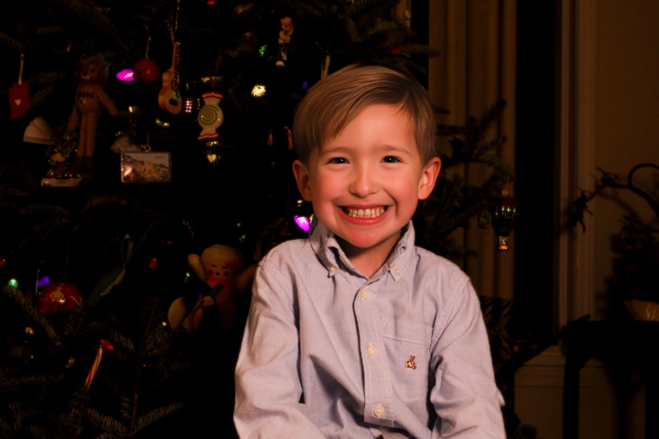 A young boy with short blond hair smiling widely, sitting in front of a decorated Christmas tree with colorful lights and ornaments, indoors.