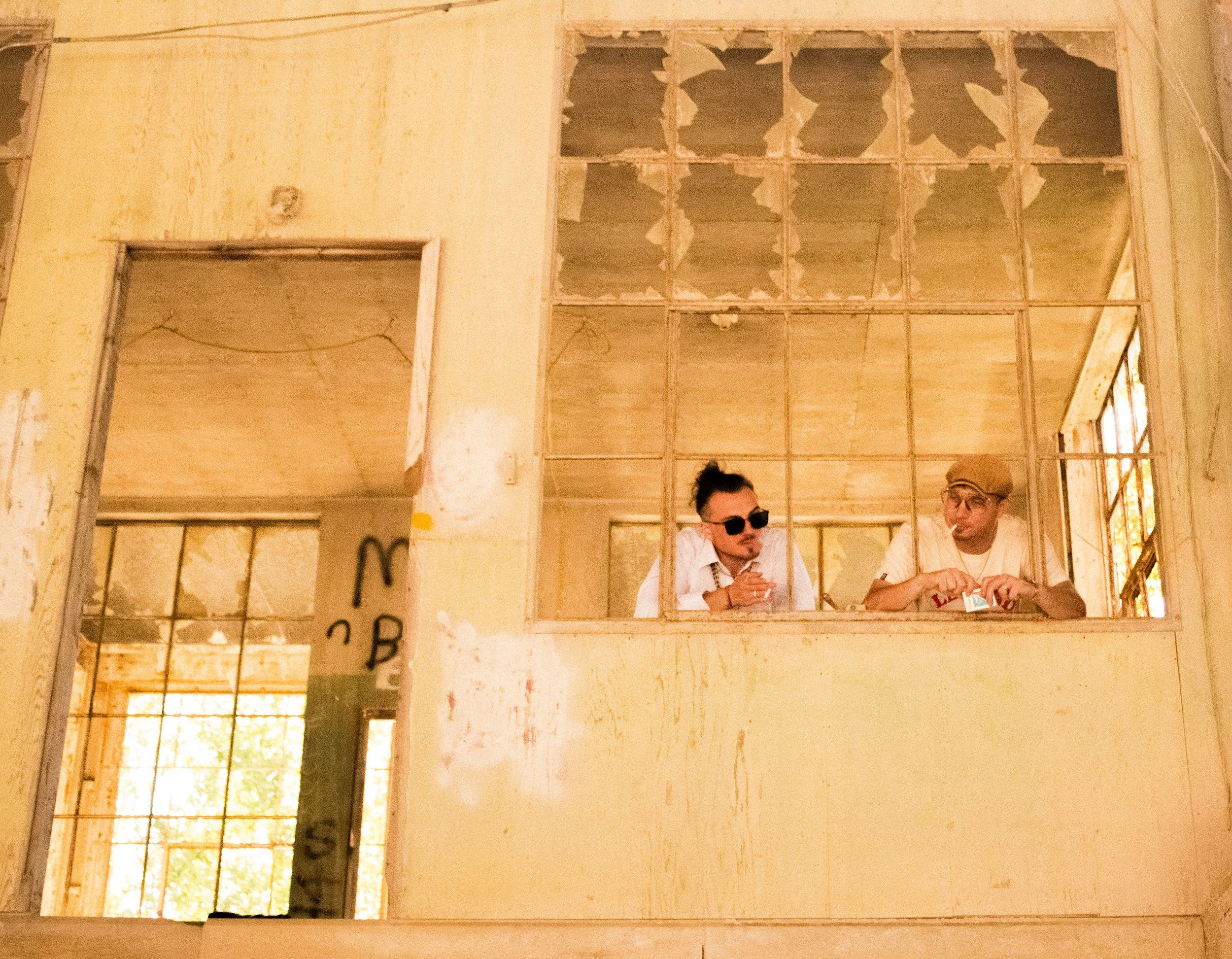 Two men working on a construction site with exposed wooden framing and a partially finished ceiling with large holes, seen through a window in a building under renovation.