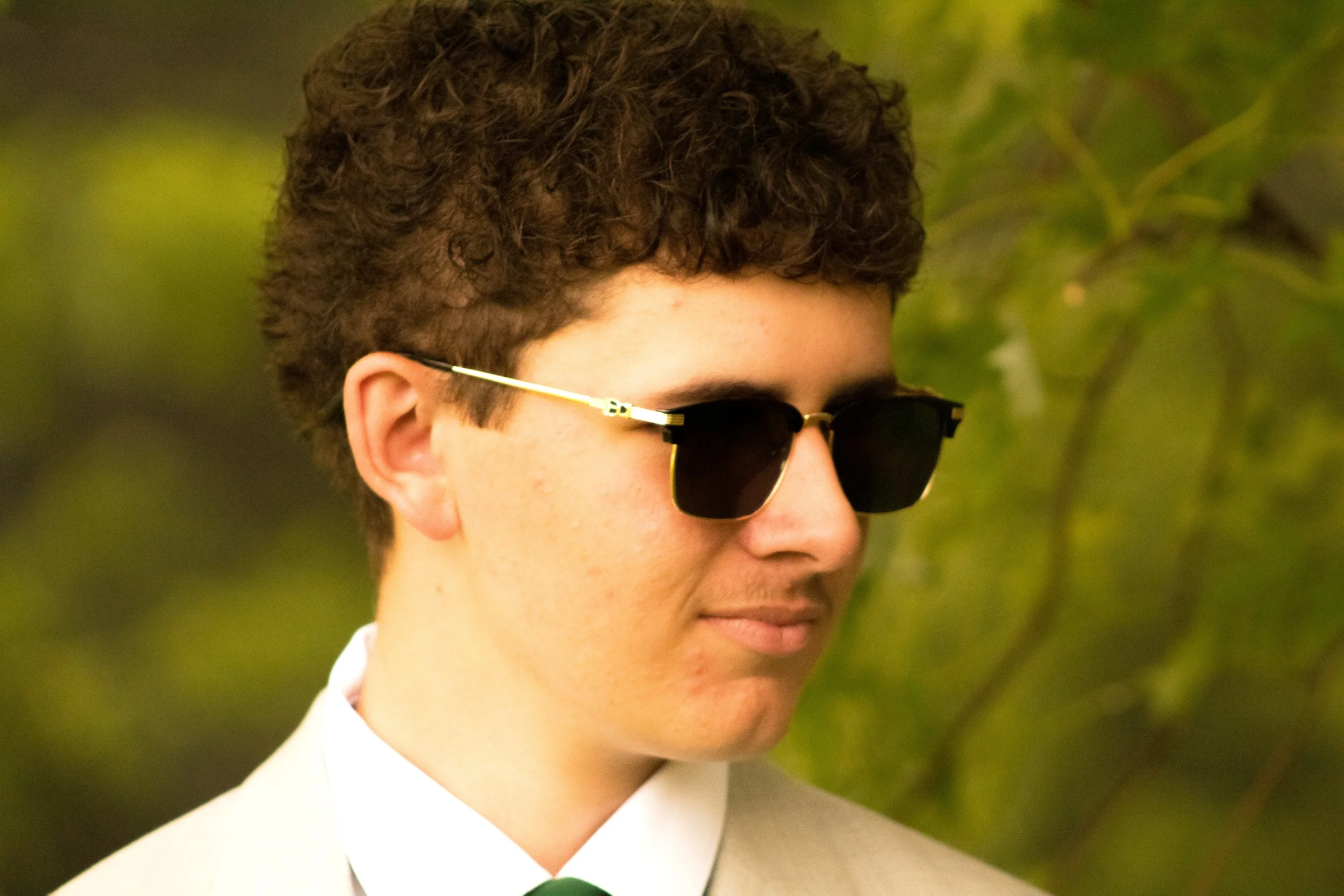 Close-up of a young man with curly hair wearing dark sunglasses and a white shirt against a blurred green background.
