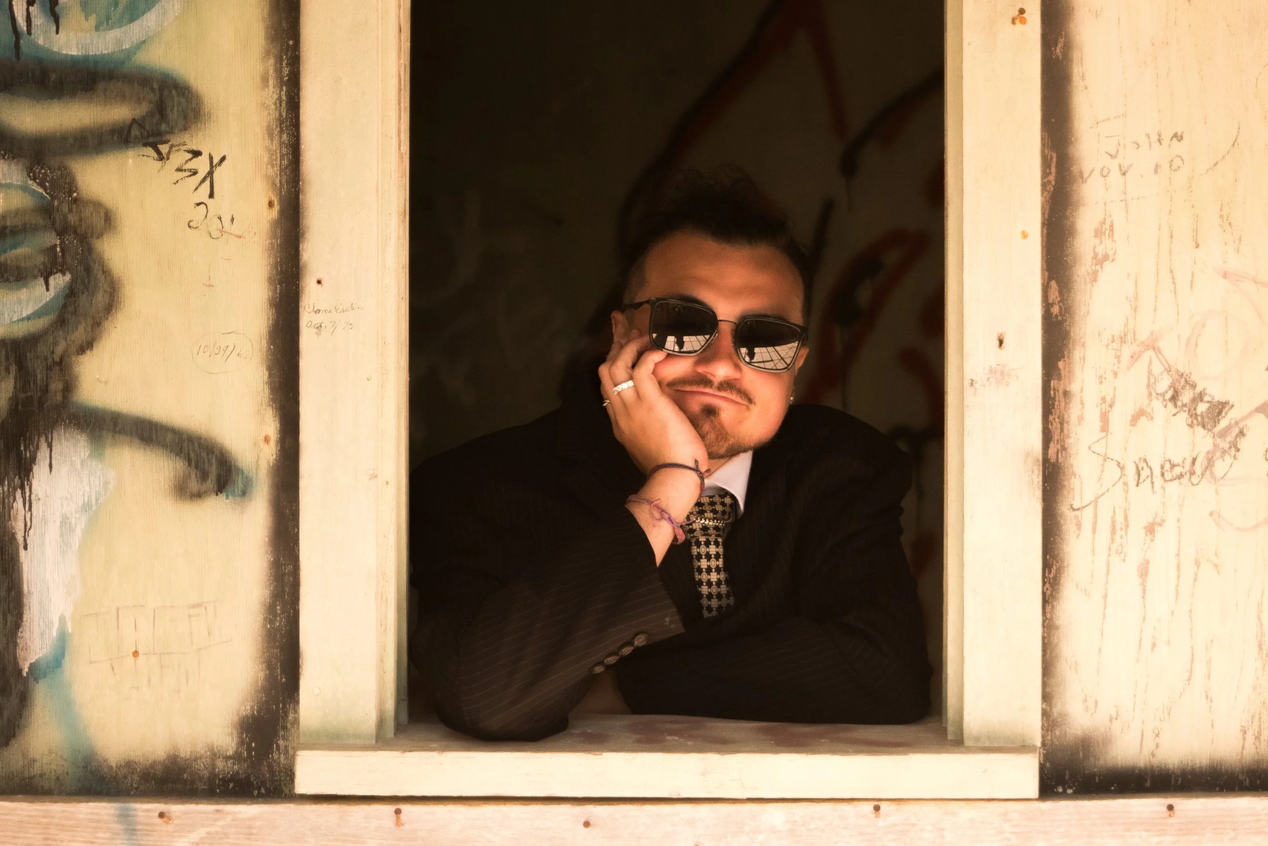 A man wearing sunglasses, a black blazer, and a patterned shirt poses with his hand on his face inside a small, graffiti-covered wooden structure with a window.