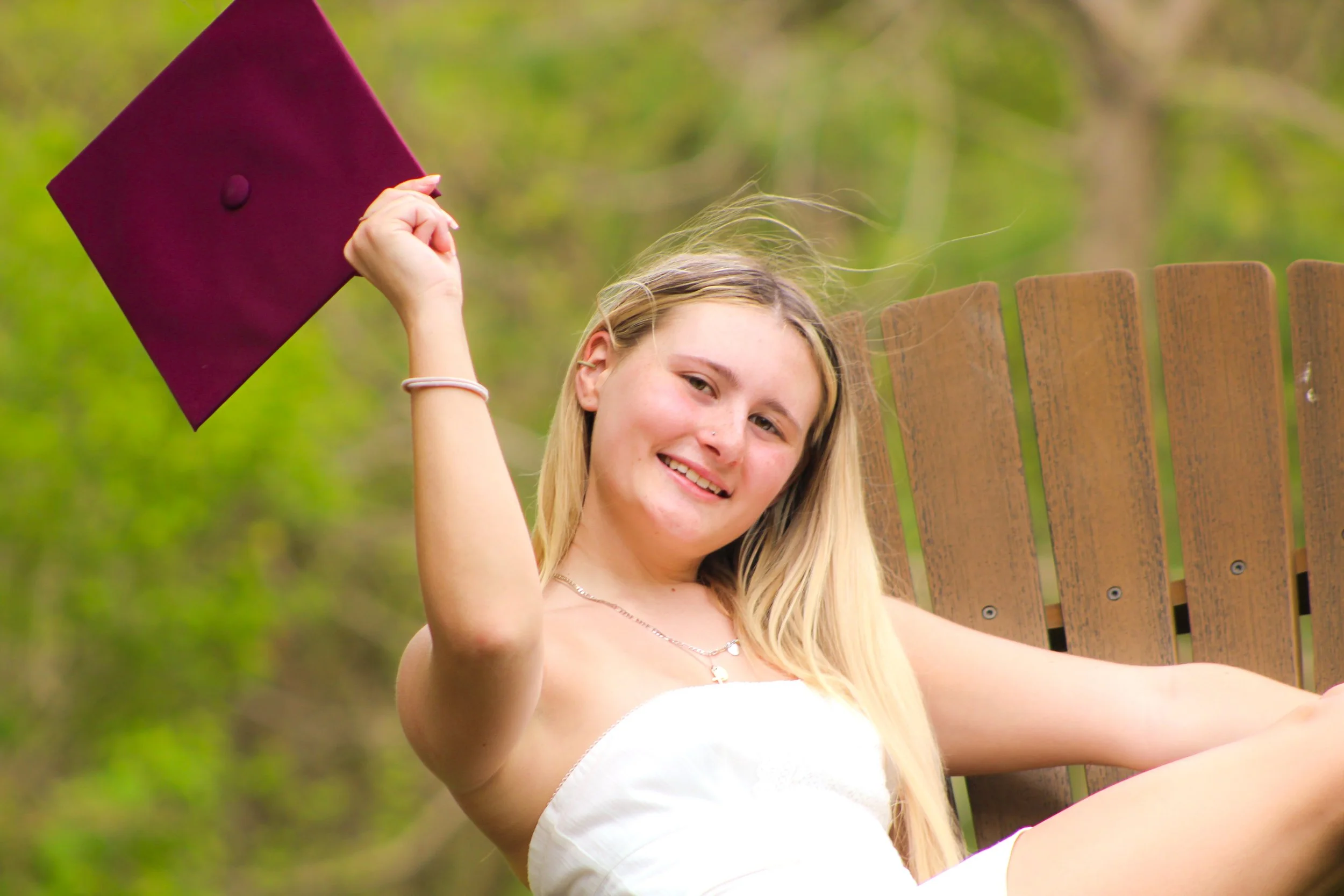 Young woman with long blonde hair smiling and holding a maroon graduation cap outdoors, sitting on a wooden chair with a background of green trees.