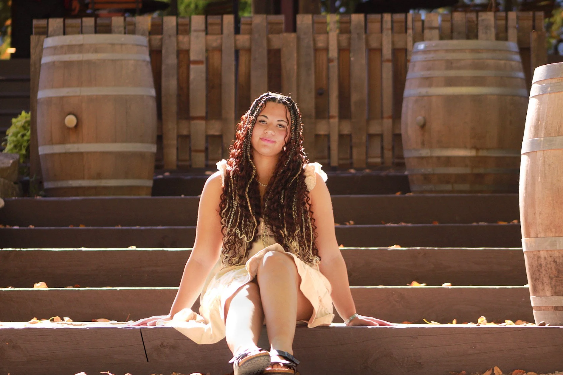 Young woman with braided hair sitting on wooden steps outdoors, surrounded by wooden barrels, in sunlight.