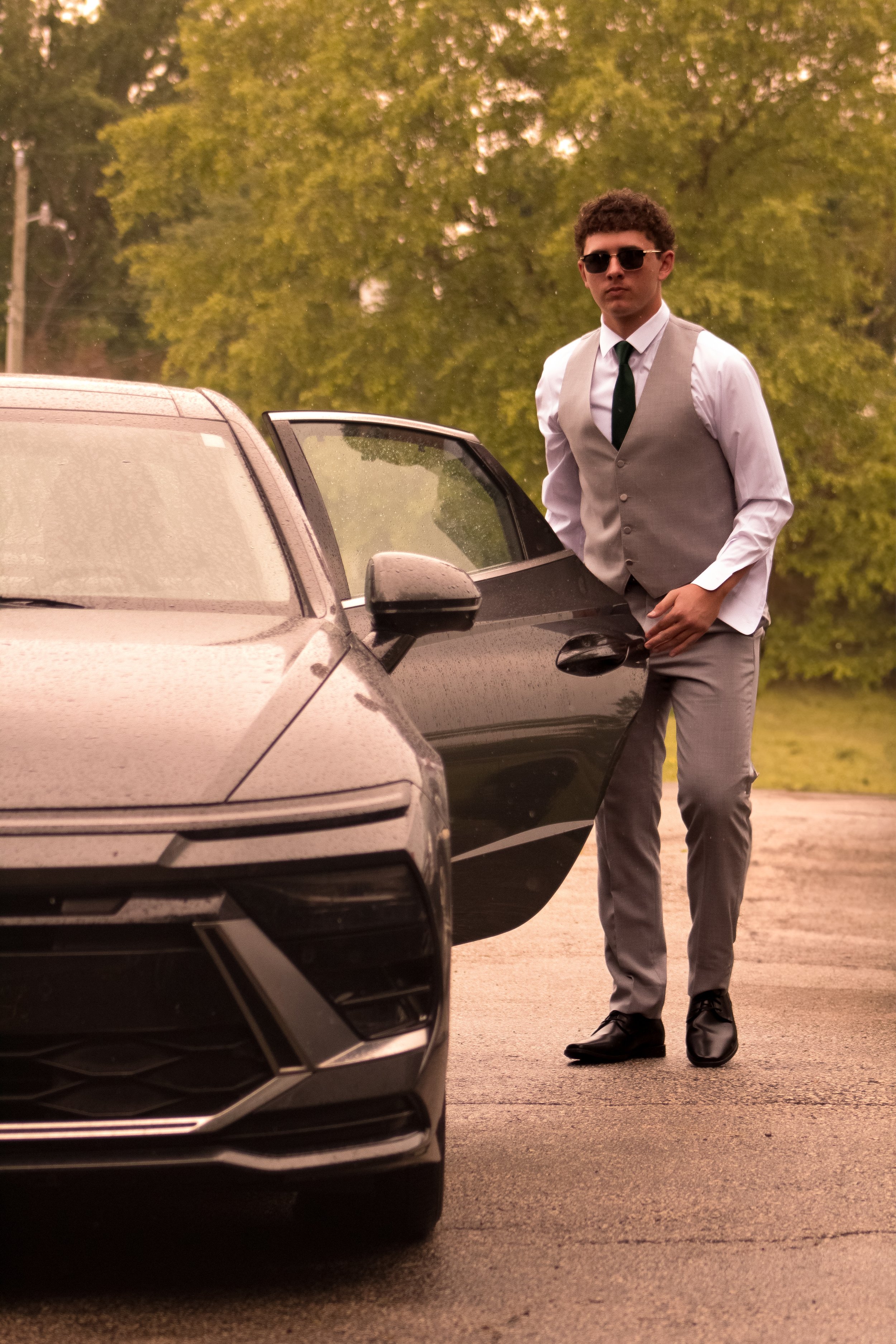 A young man wearing sunglasses, a white shirt, a gray vest, and tie, standing next to a black car with the car door open on a road with green trees in the background.
