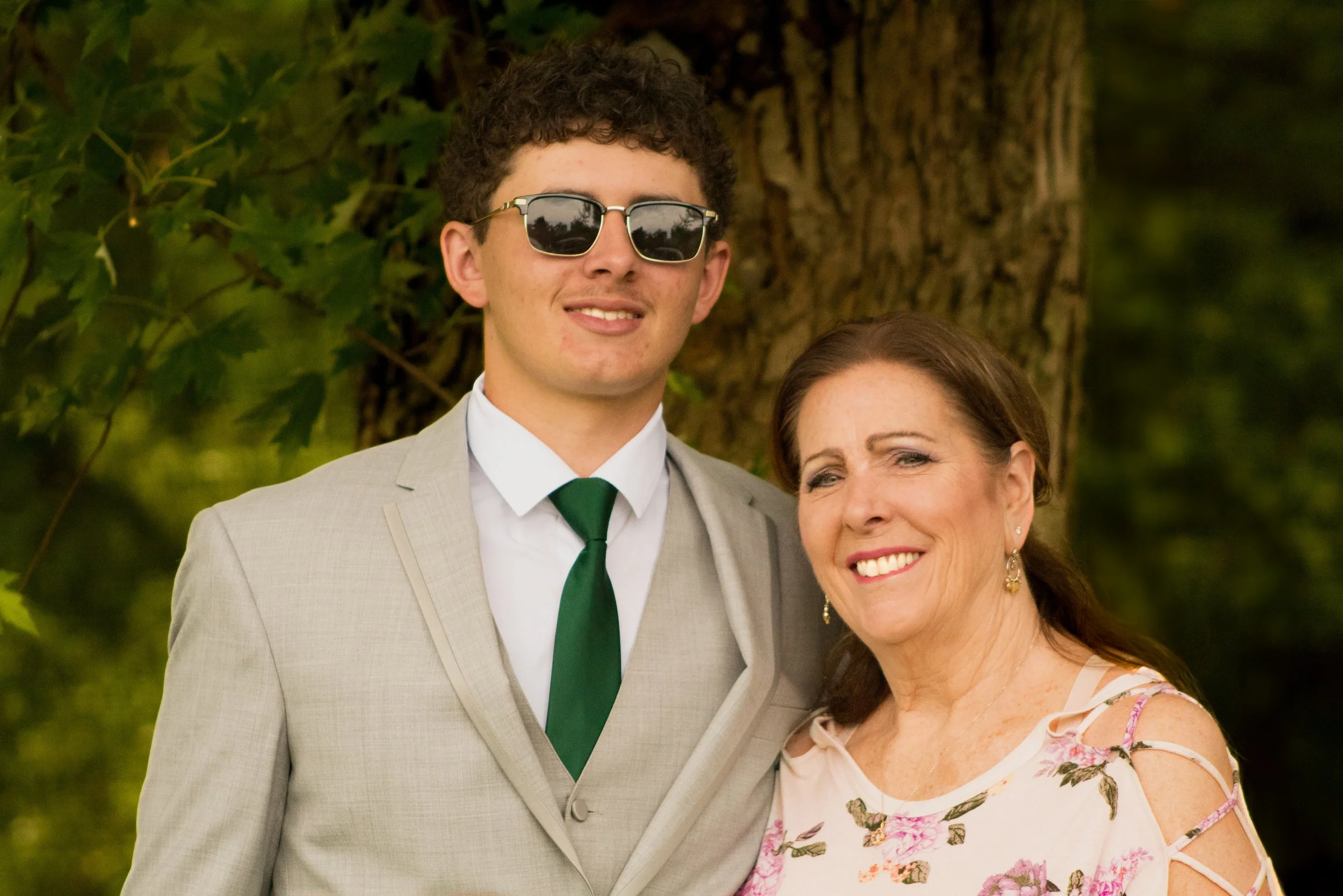 A young man in a light suit with a green tie wearing sunglasses standing next to a middle-aged woman in a floral dress smiling outdoors.