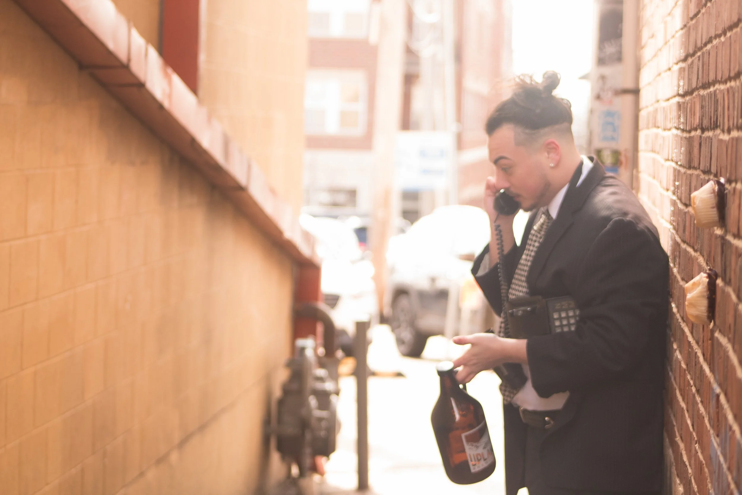 A young man in a black suit standing in a narrow alley, holding a bottle of beer and talking on a phone, with brick walls on either side and sunlight in the background.