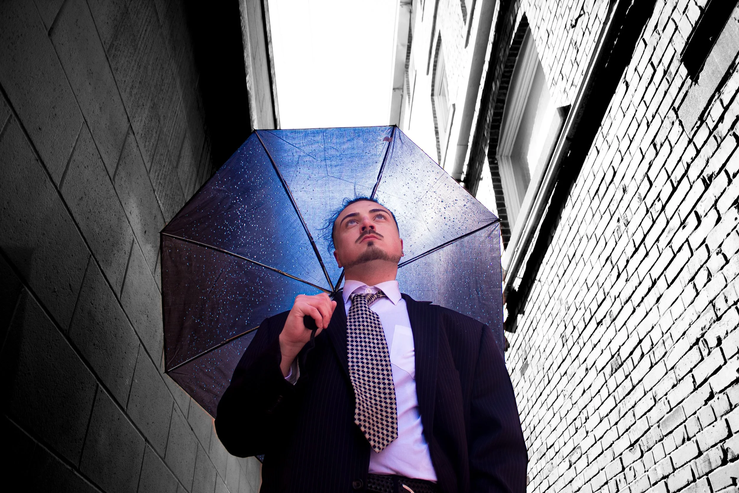 Man in a suit holding an umbrella, standing in a narrow alleyway between brick and concrete buildings on a rainy day.